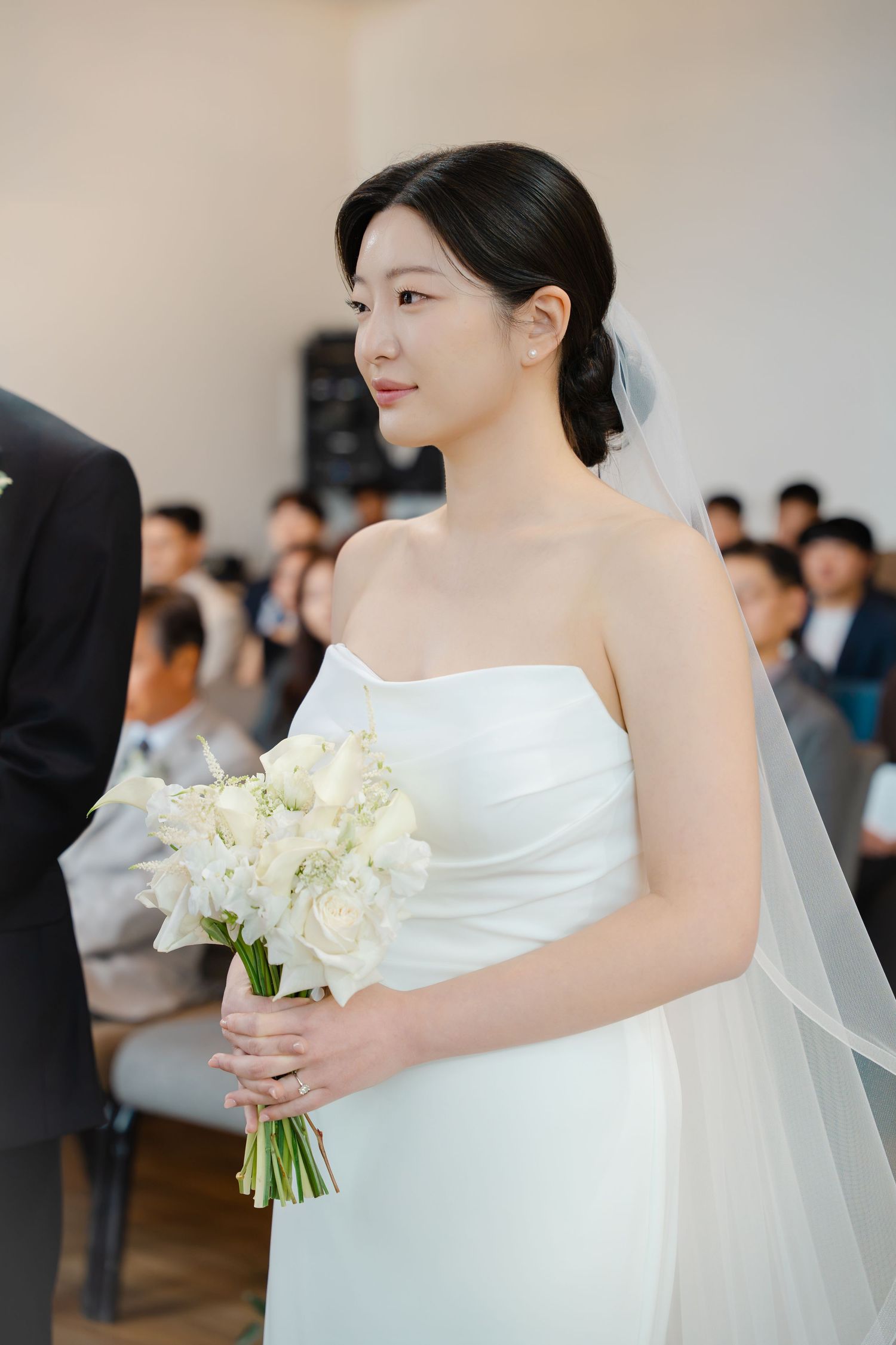 Elegant bride in strapless white gown holds white calla lily bouquet during indoor wedding ceremony.