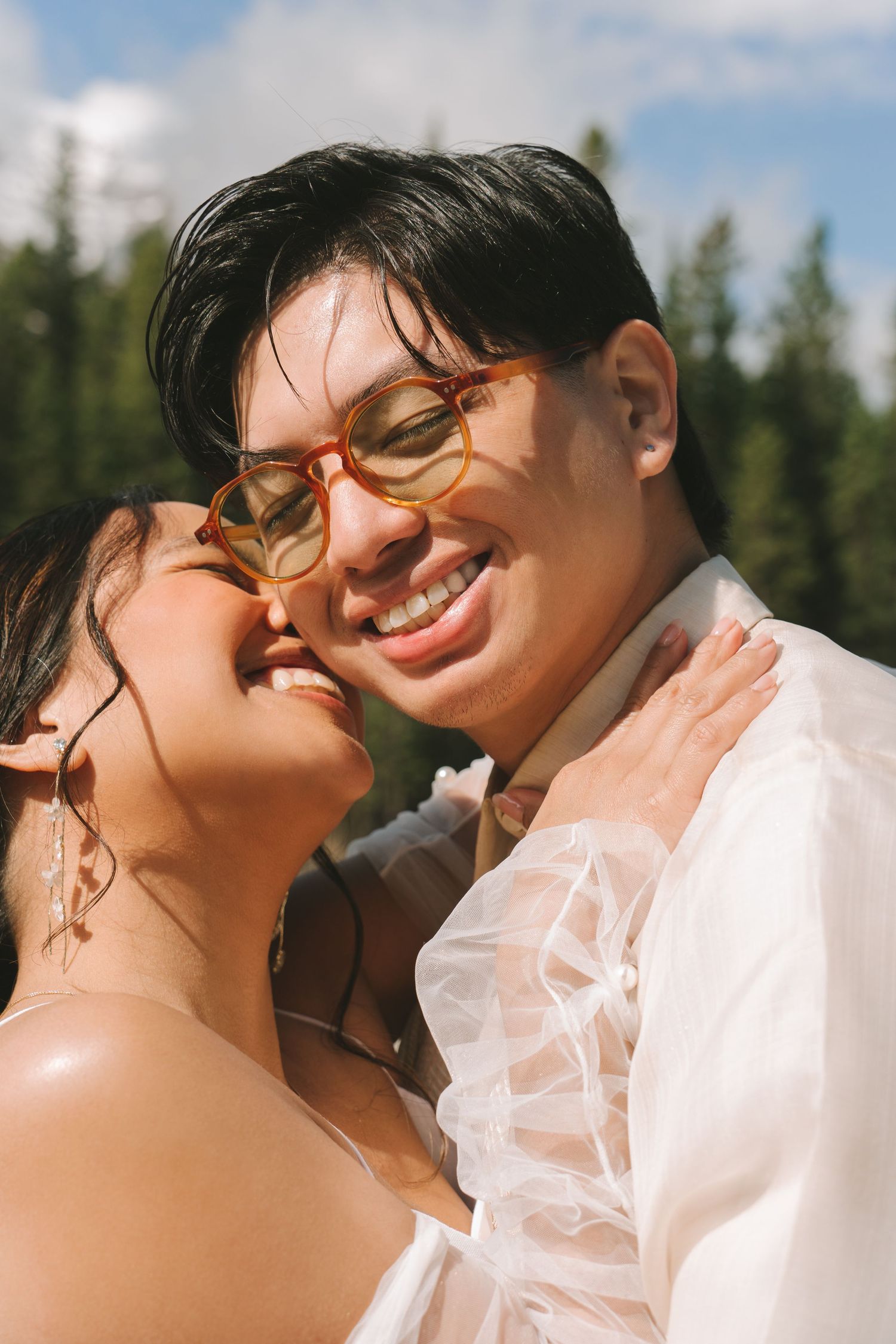 Smiling couple in wedding attire embrace outdoors with pine trees and blue sky in background.