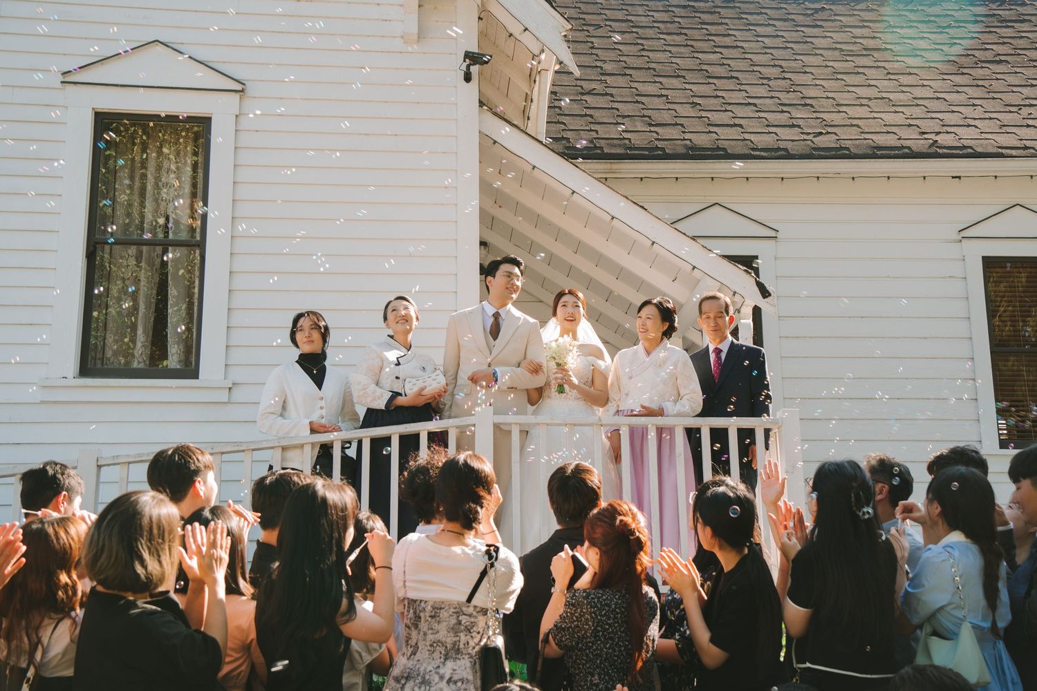 Wedding party celebrates on white building balcony as guests below toss confetti in joyful outdoor ceremony moment.