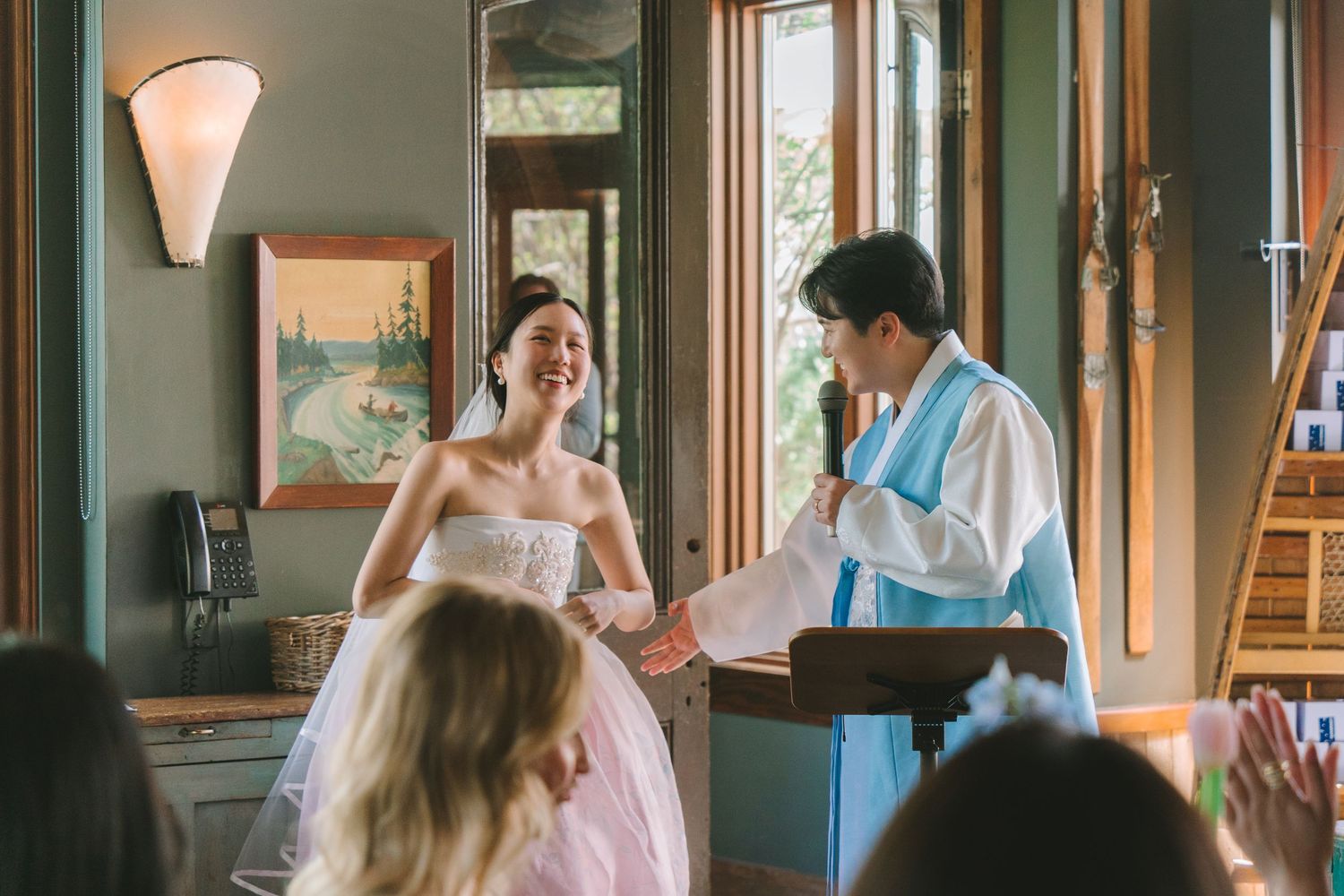 Laughing Asian bride in pink gown holds hands with groom in blue hanbok during wedding reception.
