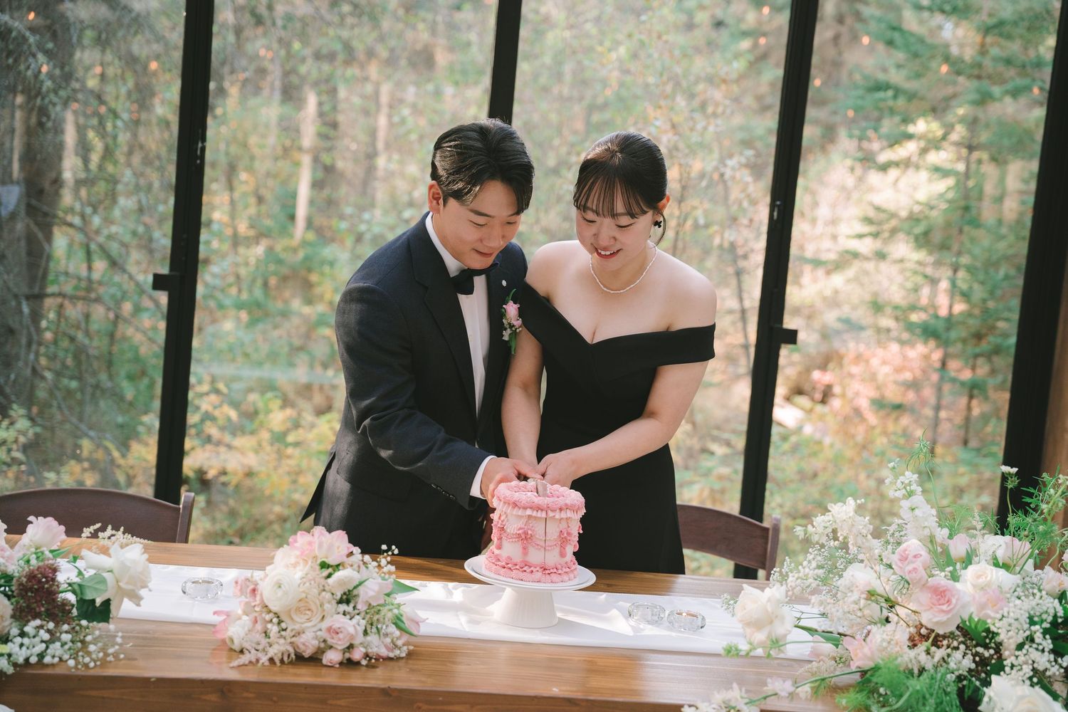 Asian couple cuts pink wedding cake at a table decorated with white flowers in a glass-walled venue.