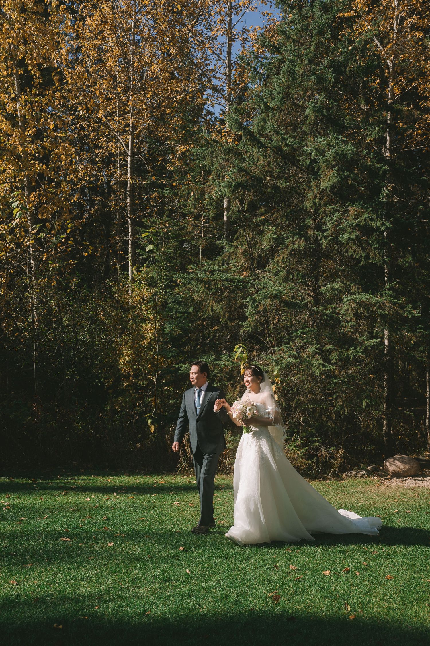 Bride and groom walk hand in hand across a sunny green lawn surrounded by colorful autumn trees.