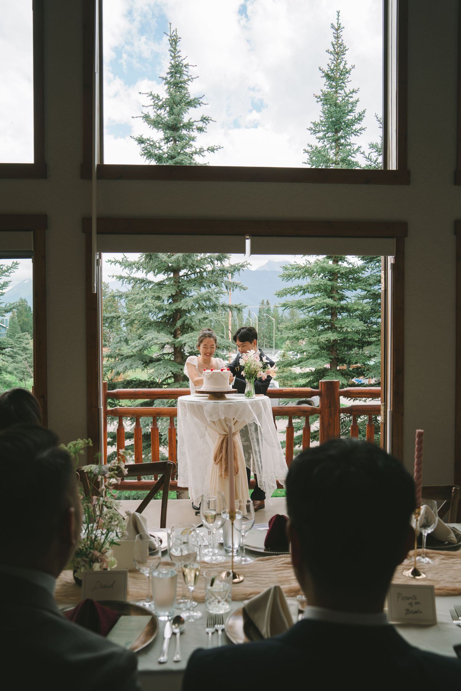 Bride and groom cutting cake by large window overlooking pine trees.