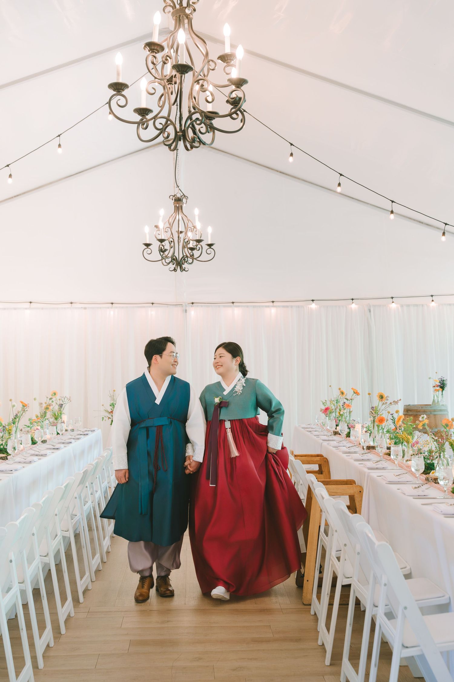 Korean couple in traditional hanbok walking hand in hand through an elegant tent wedding reception venue.