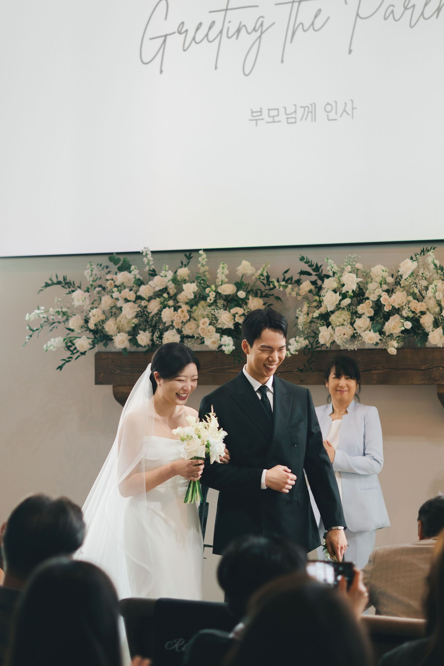 Bride and groom walk together smiling at Korean wedding reception beneath greeting the parents sign.