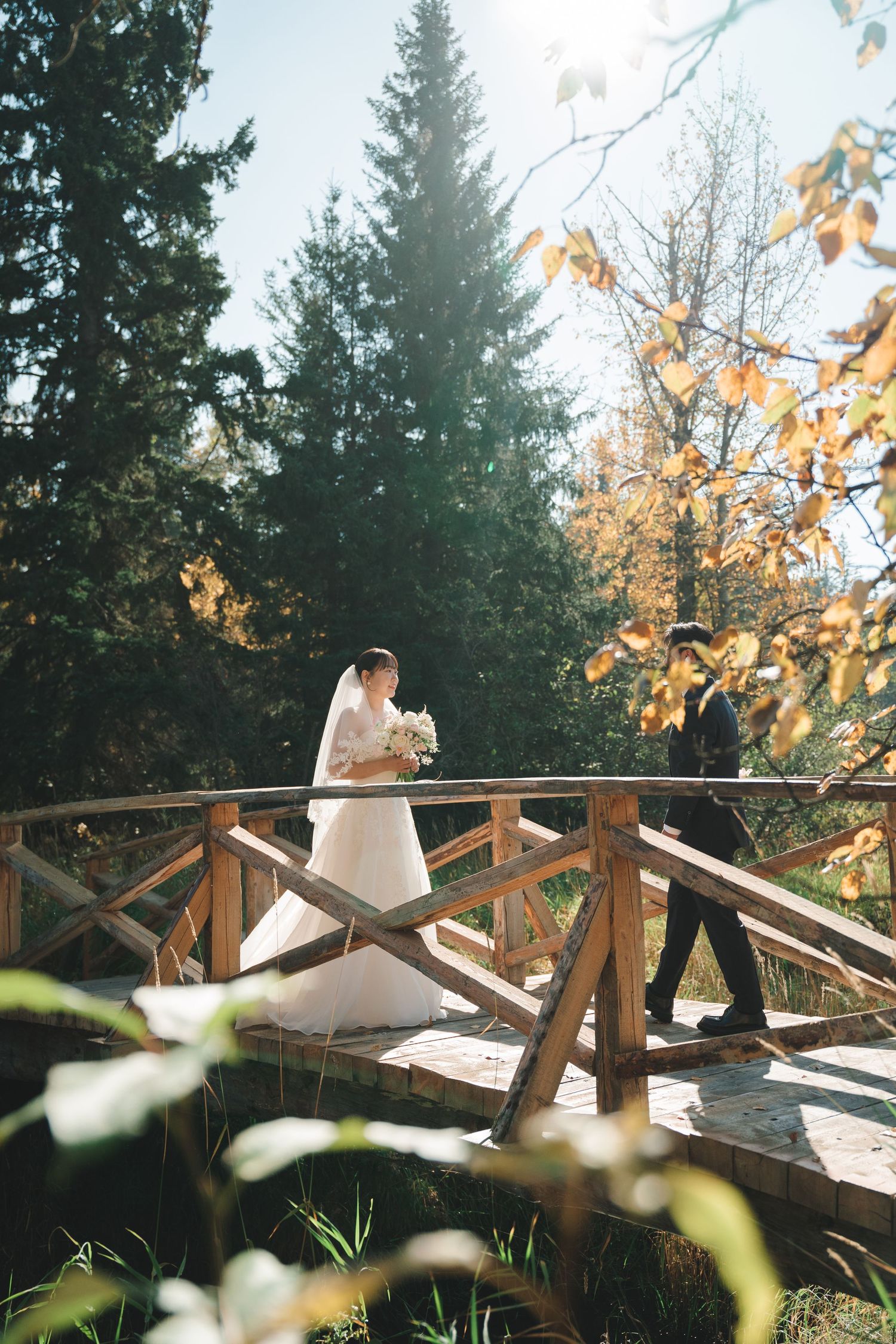 Wedding couple strolls across a rustic wooden bridge amid golden autumn foliage and tall evergreen trees.