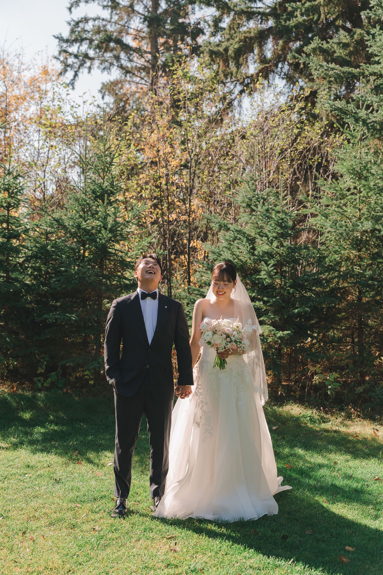 Bride and groom hold hands while strolling on green grass with evergreen trees in the background.