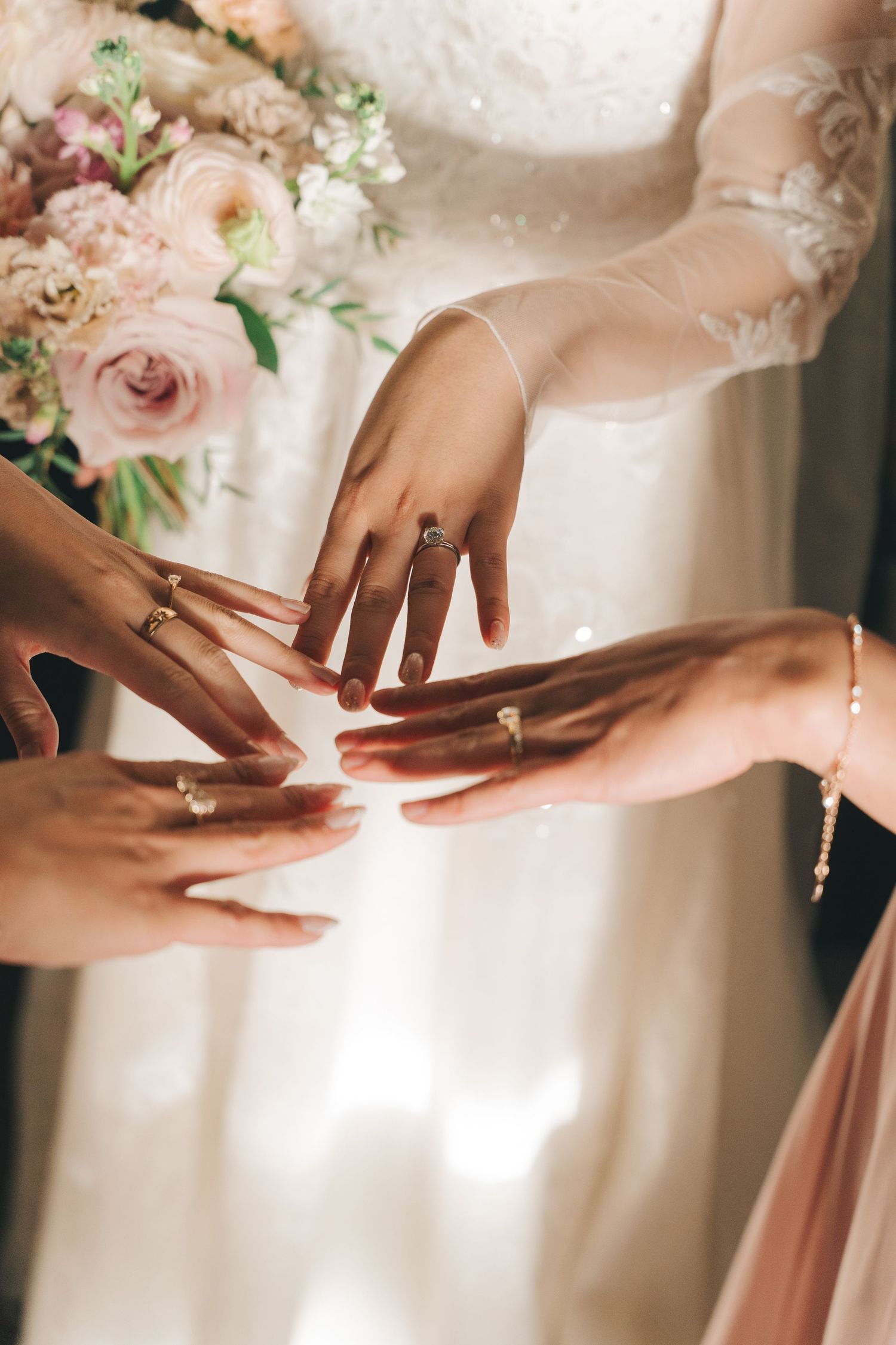 Bride and bridesmaids touch hands together showing their rings near a soft floral bouquet.