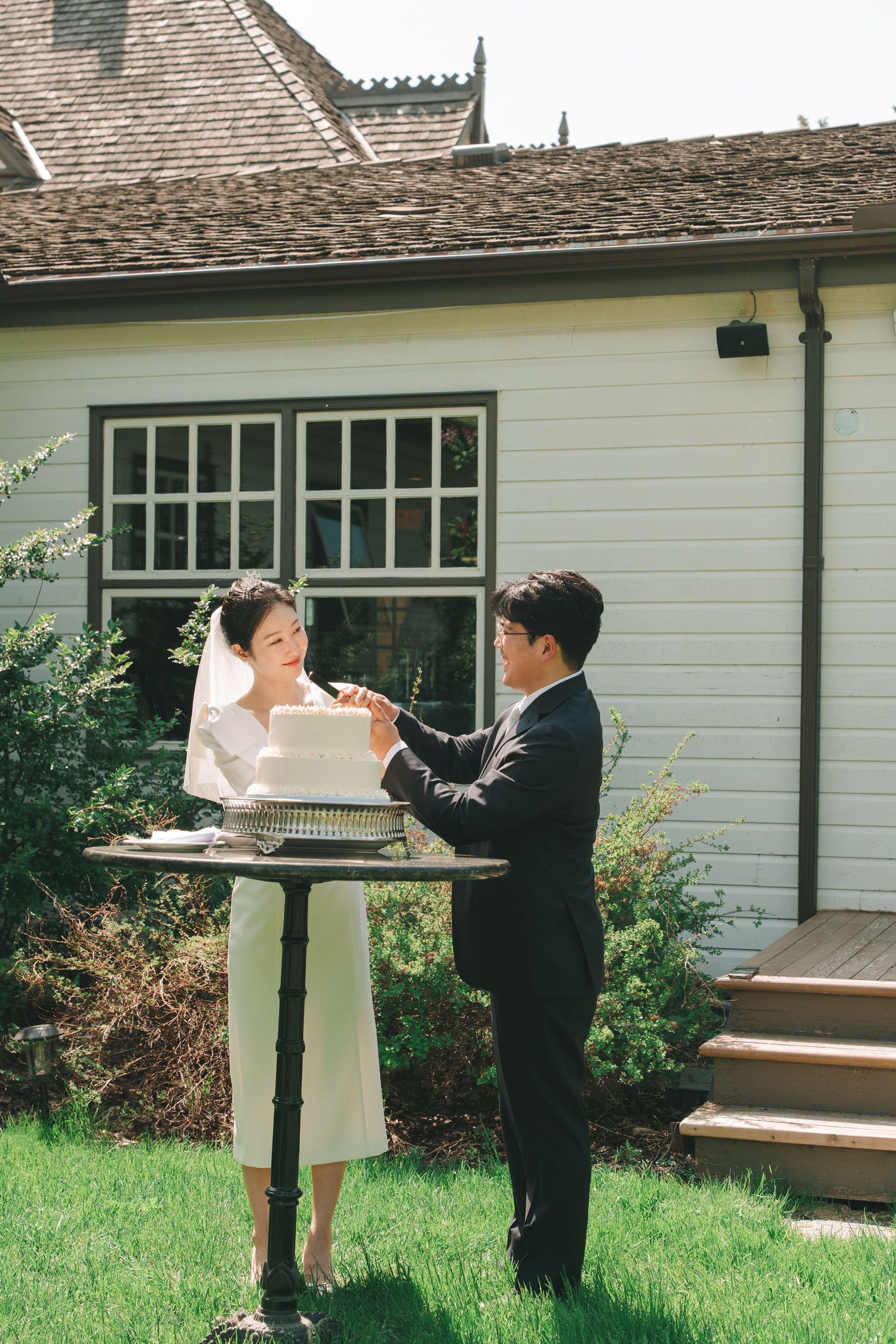 Bride and groom share cake cutting moment in sunny garden beside white historic building.