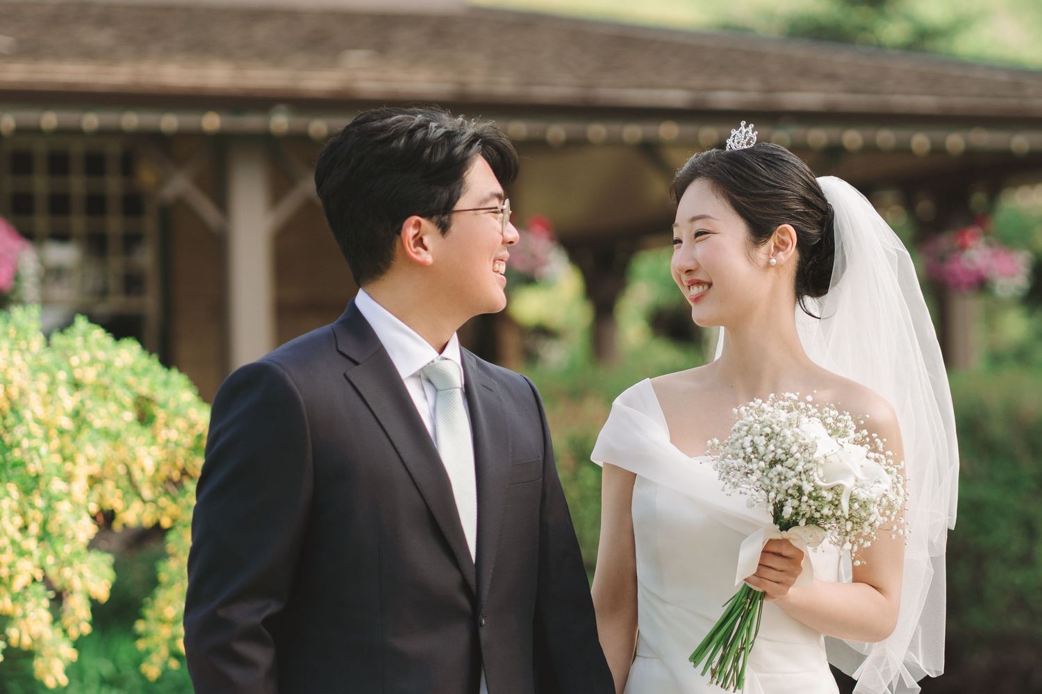 Asian bride holding baby's breath bouquet smiles at groom in lush garden setting.
