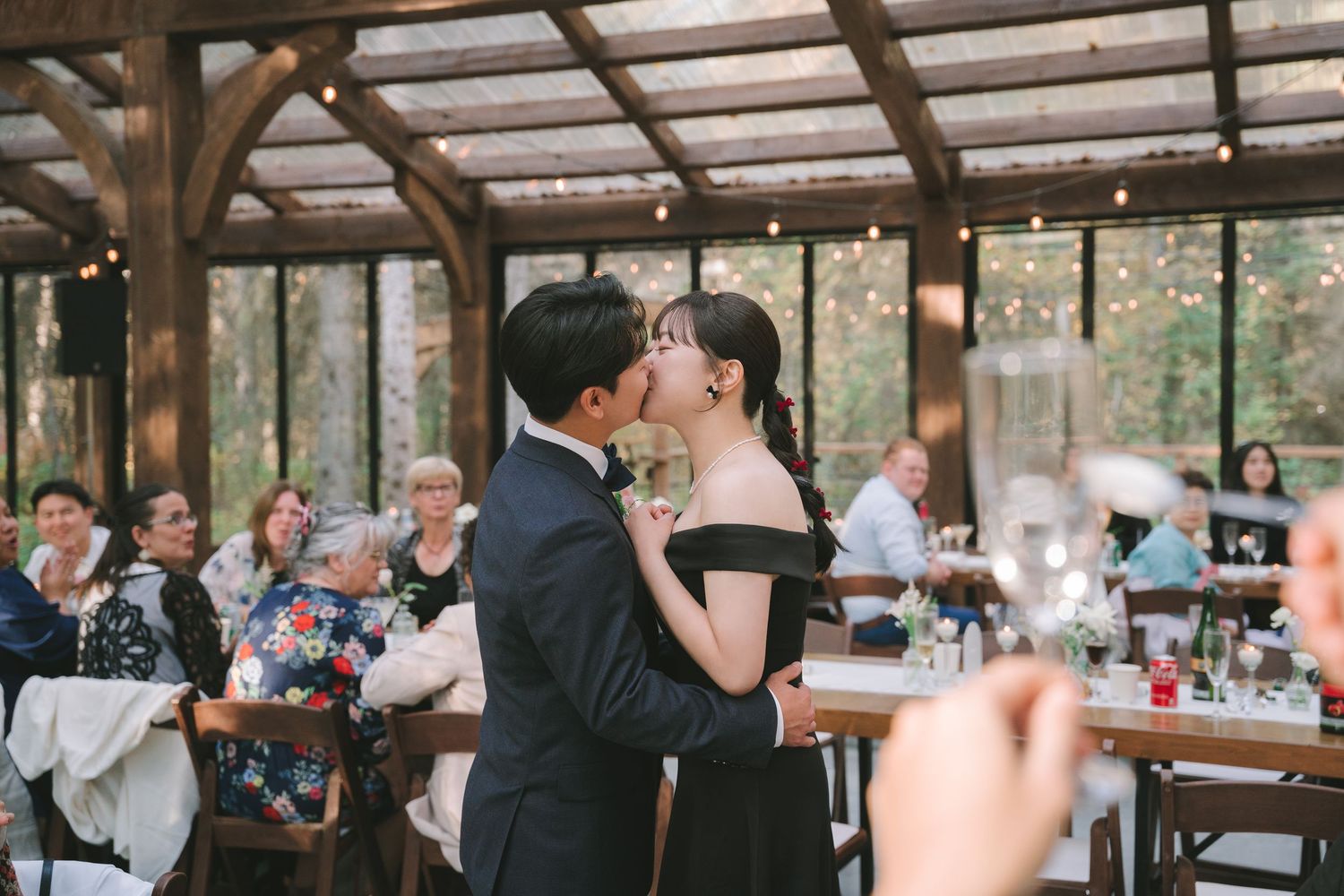 Asian couple shares first dance kiss in a warmly lit rustic reception hall surrounded by wedding guests.