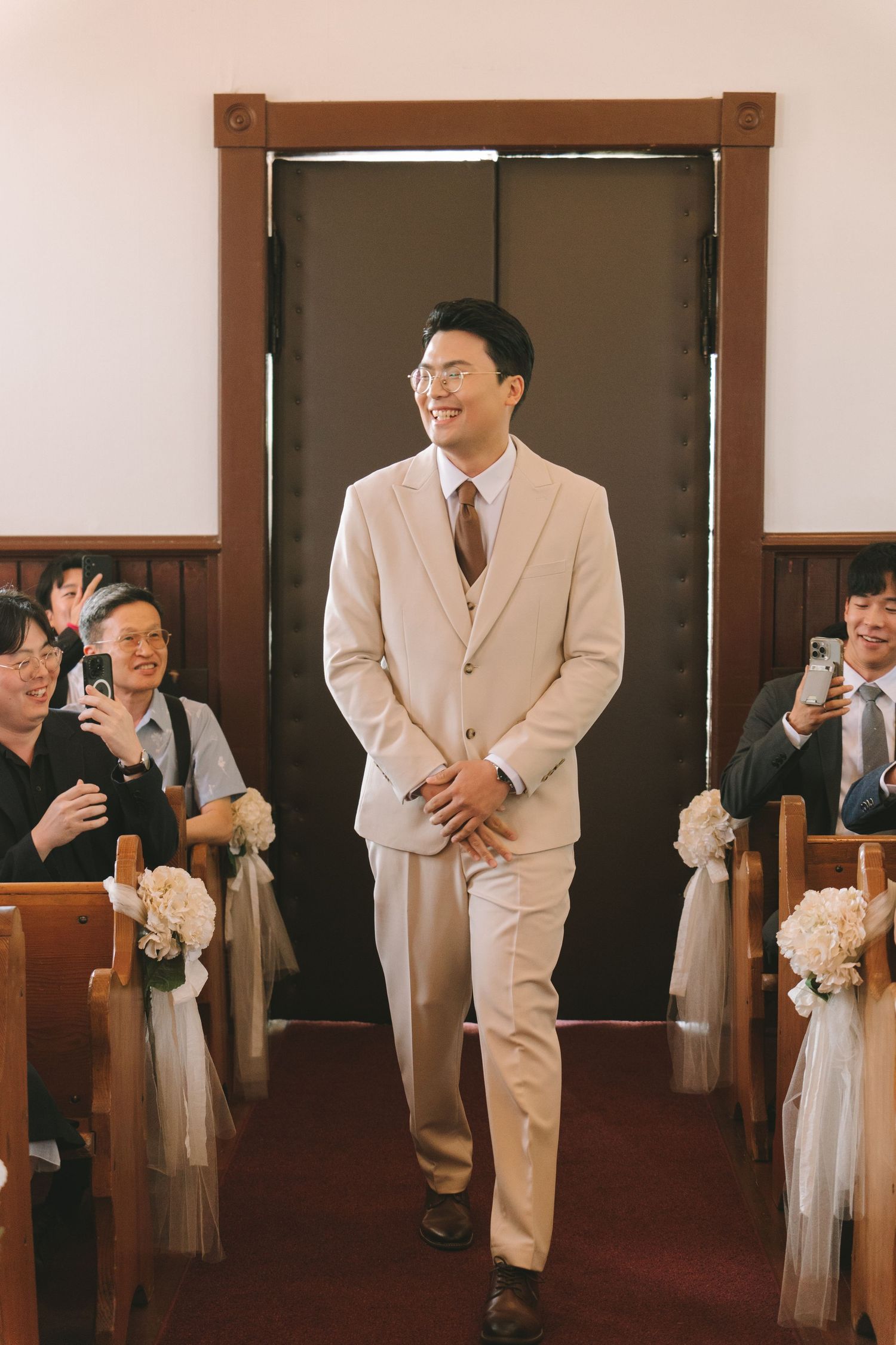 Smiling groom in beige three-piece suit walks down church aisle lined with floral decorations.