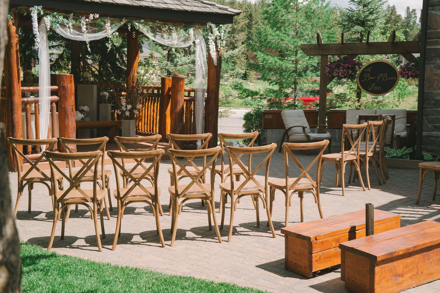 Rustic outdoor wedding ceremony setup with wooden crossback chairs and pergola surrounded by lush greenery.