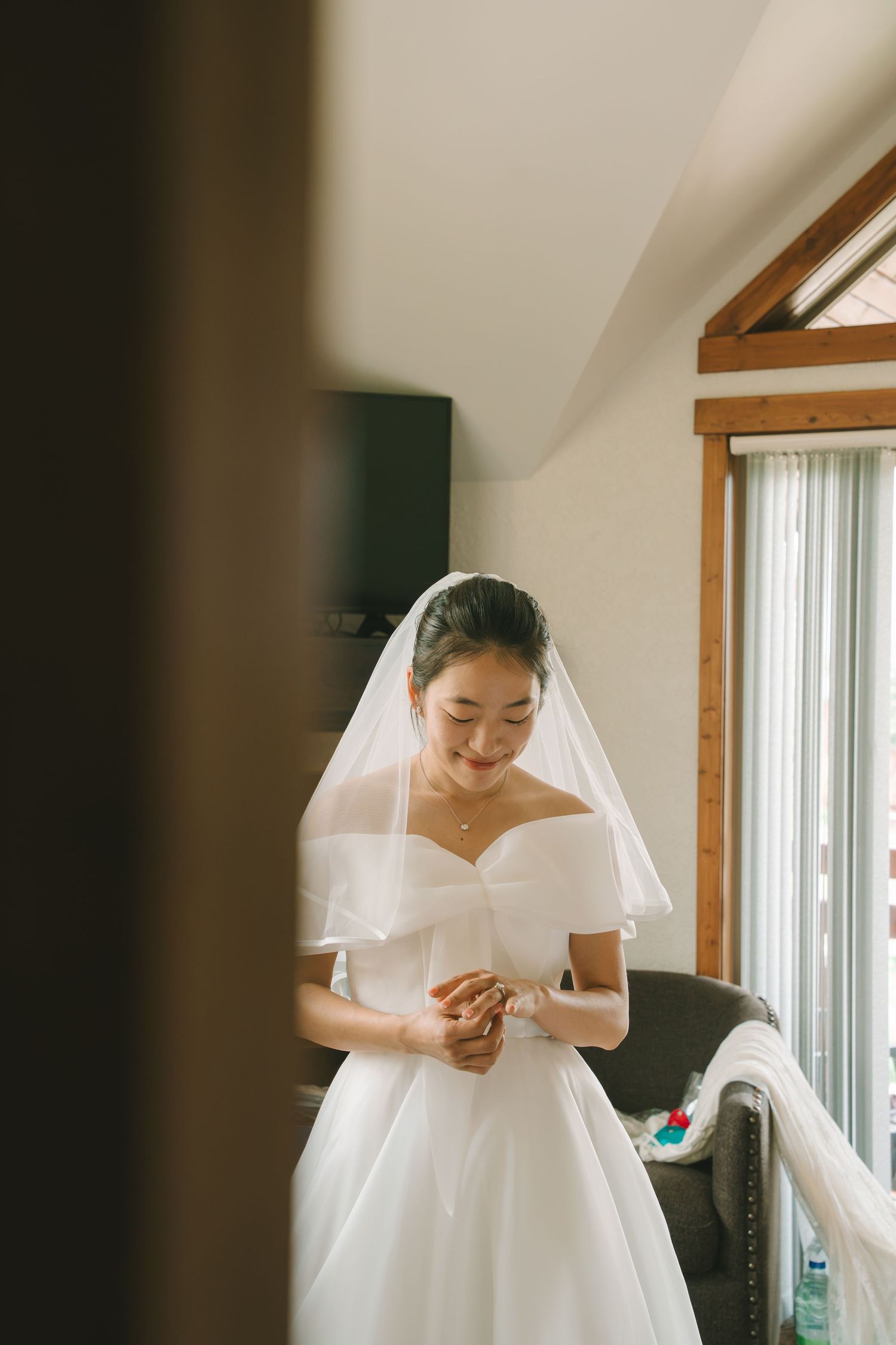 Bride in elegant off-shoulder white gown and veil looks down while getting ready in a sunlit room.