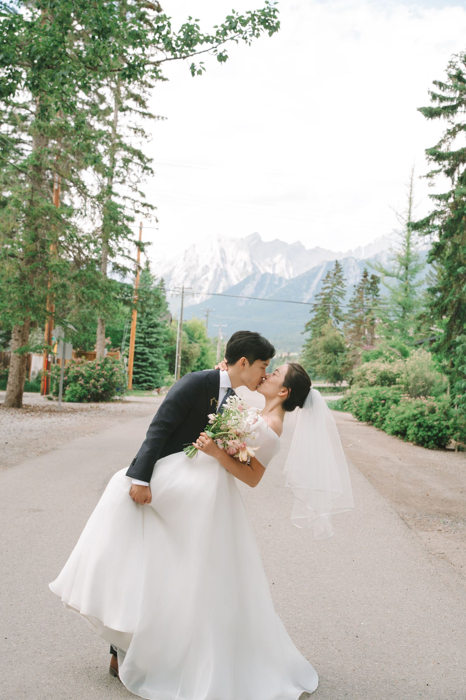Groom dipping bride for a kiss on a tree-lined path with stunning snowy mountain backdrop.