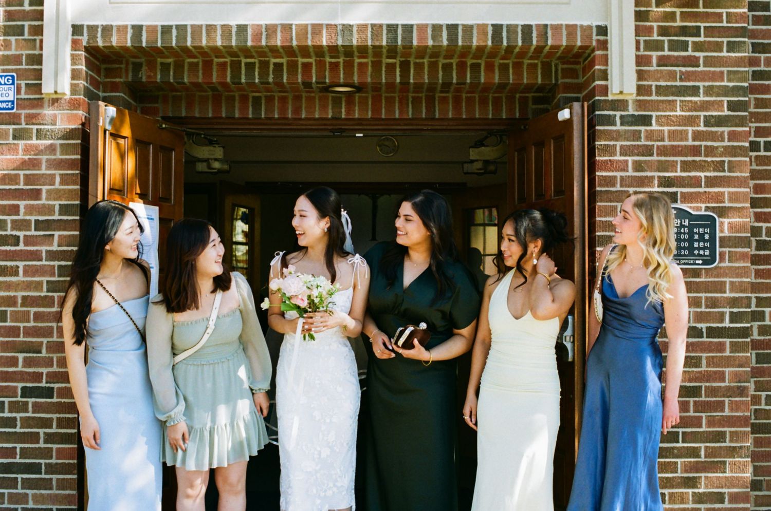 Bride in white dress laughs with five bridesmaids in varied dresses outside brick building doorway.