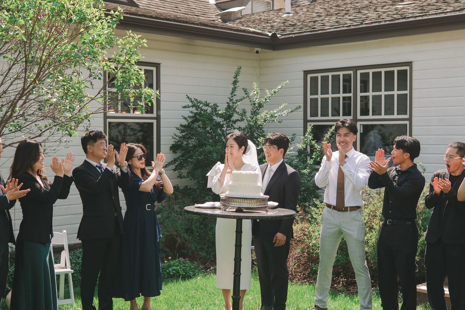 Wedding party applauds as bride and groom cut cake in charming outdoor garden venue.