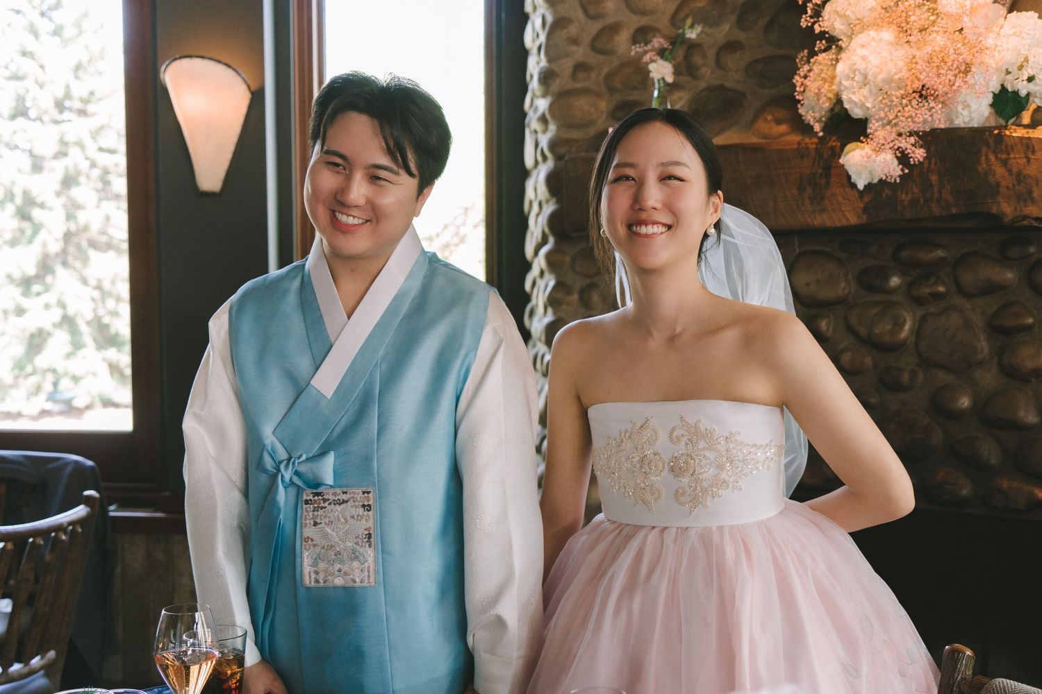 Happy Asian couple smiles at wedding reception, groom in blue hanbok and bride in pink strapless gown.