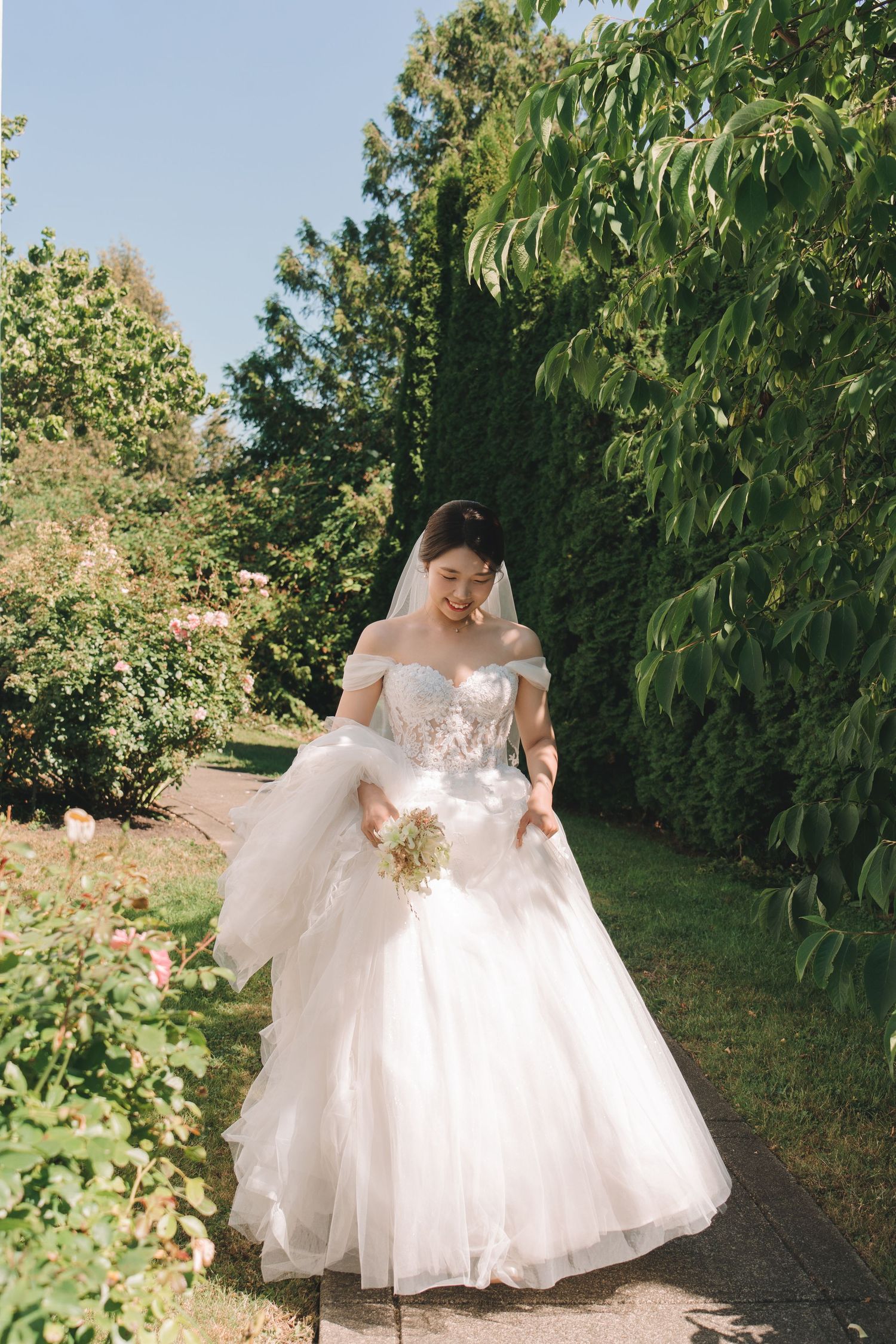 Bride in off-shoulder white ballgown walking through garden on sunny day.