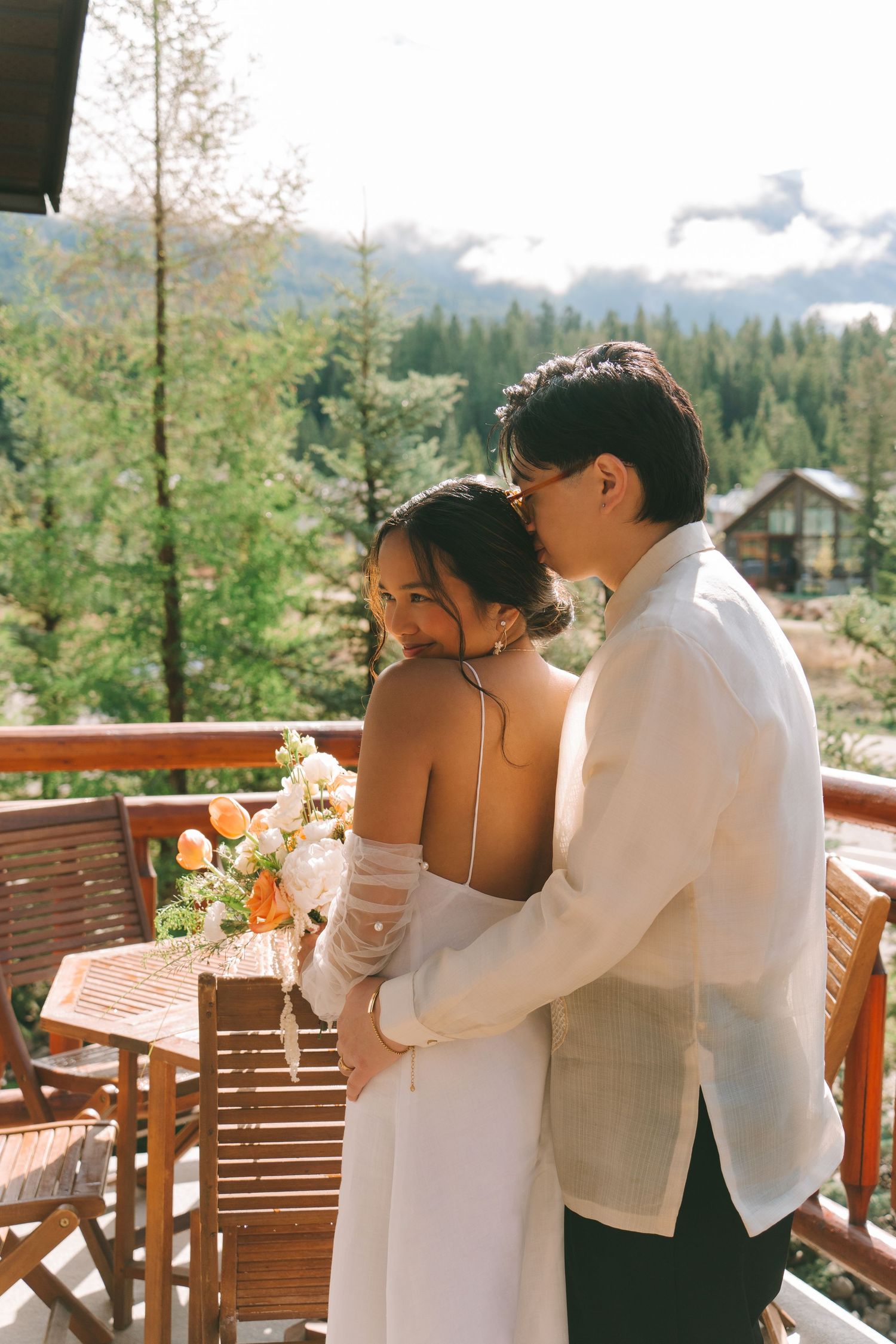 Groom embraces bride from behind on rustic balcony with mountain and pine forest views.