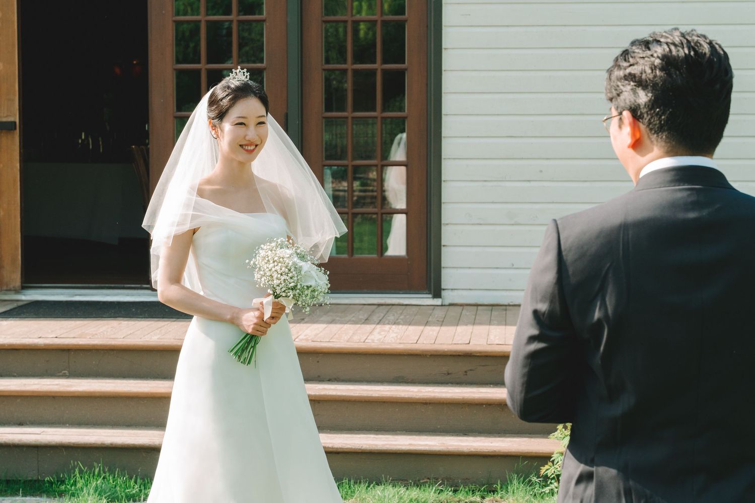 Smiling bride in off-shoulder gown holds baby's breath bouquet during first look with groom.