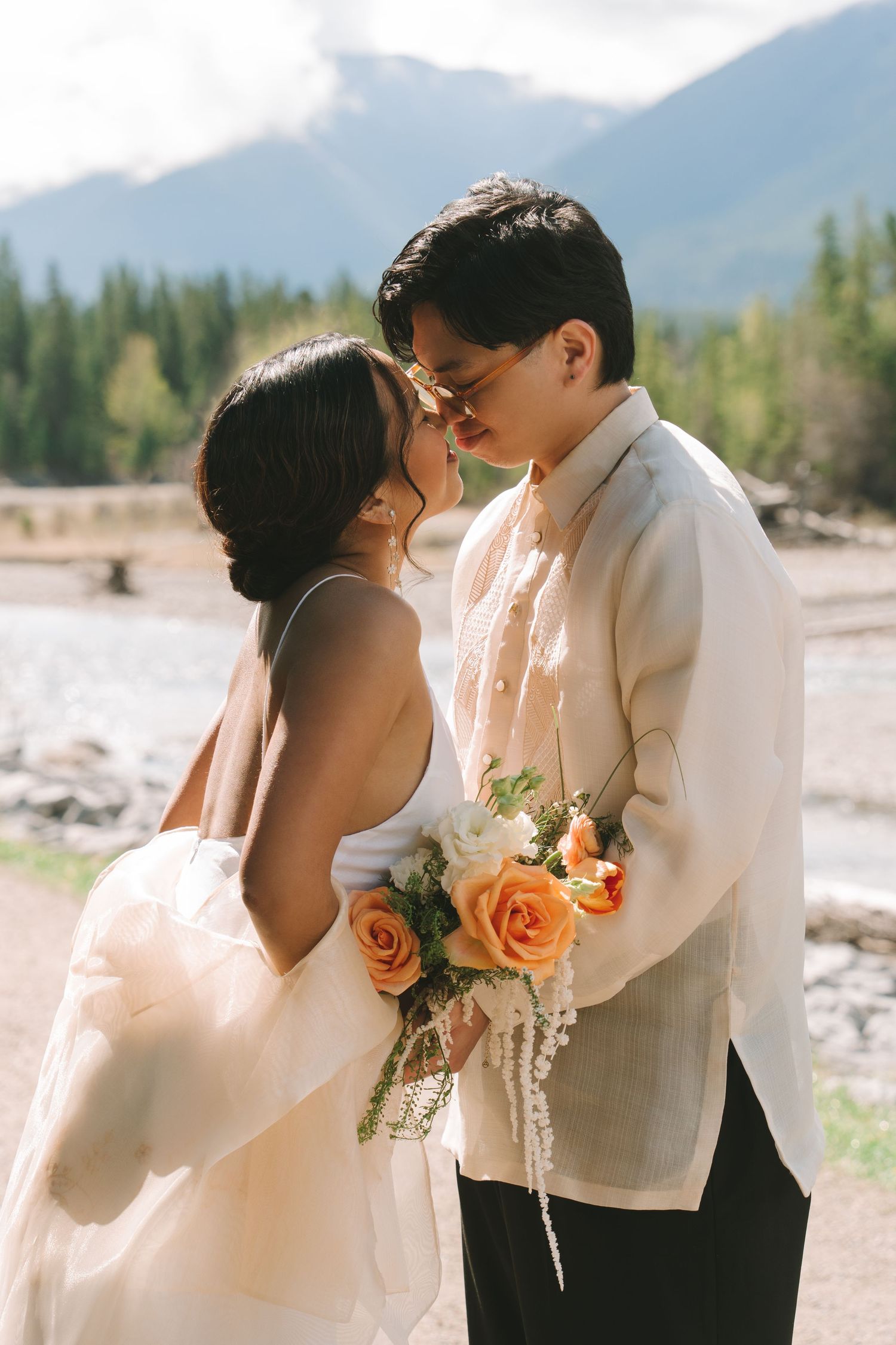Bride and groom share a romantic moment with orange roses bouquet, mountains and pine trees in background.