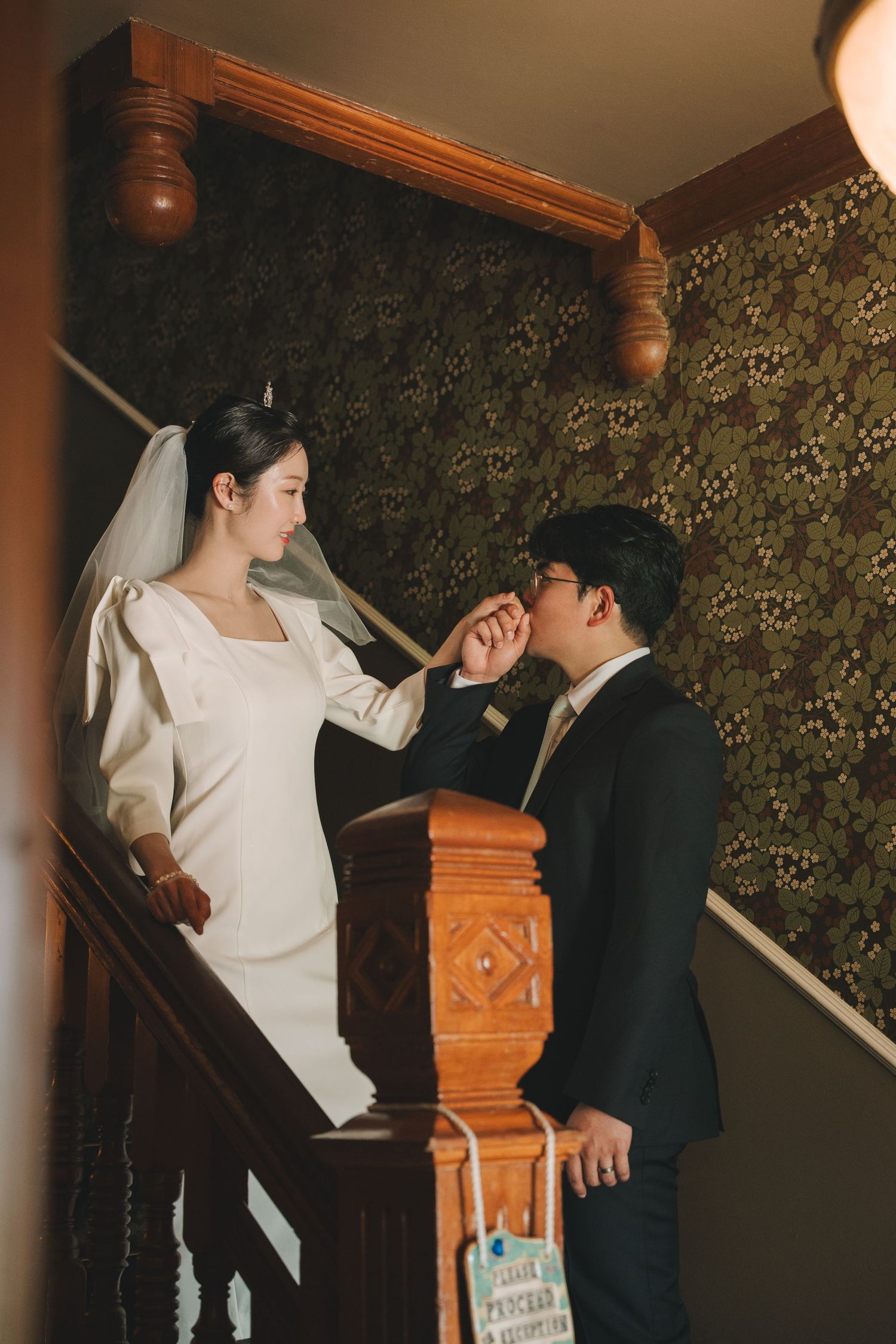 Groom kissing bride's hand on elegant wooden staircase with dark floral wallpaper backdrop.