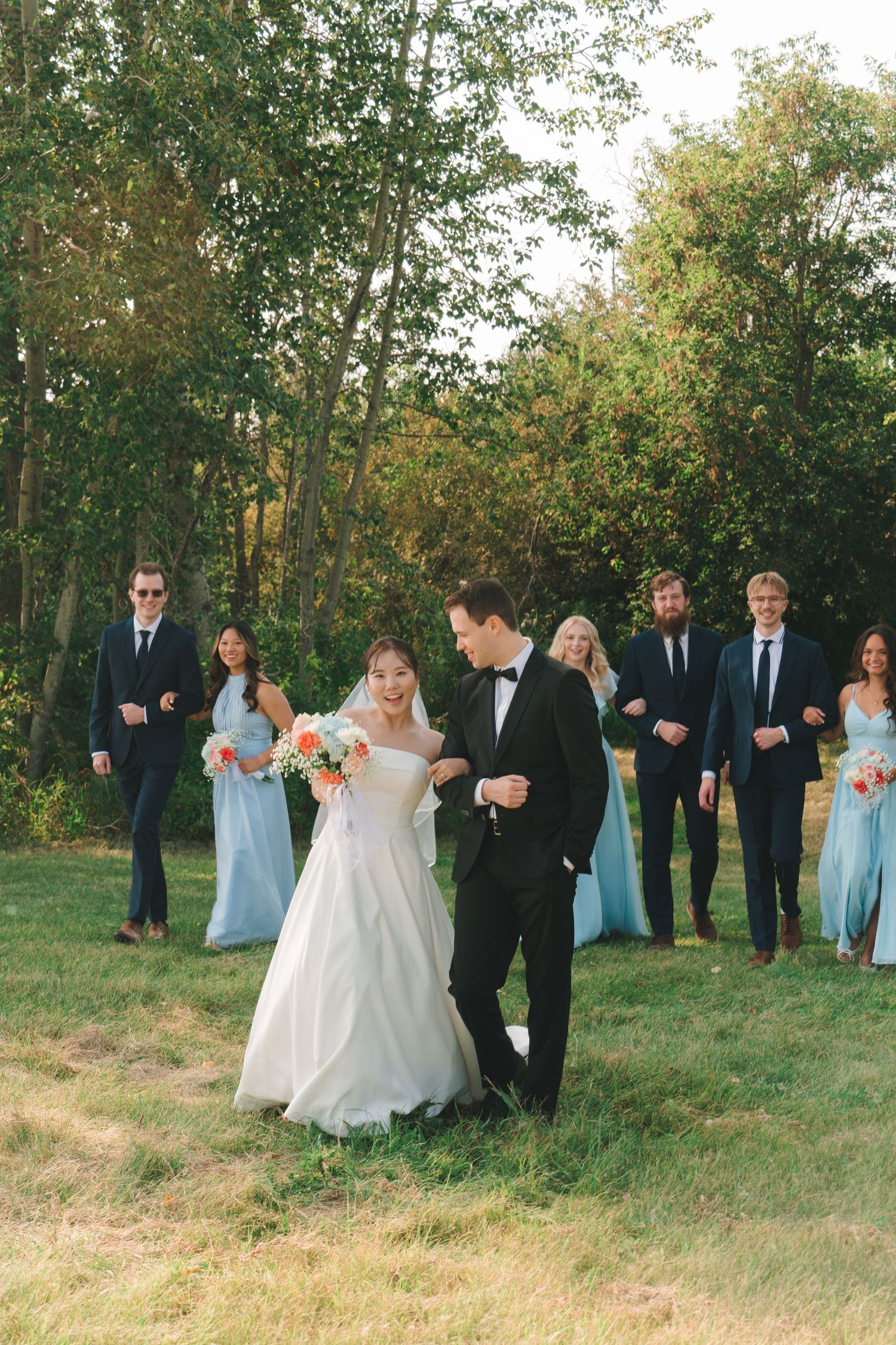 Bride and groom walk together on grass while wedding party follows behind in blue dresses and dark suits.