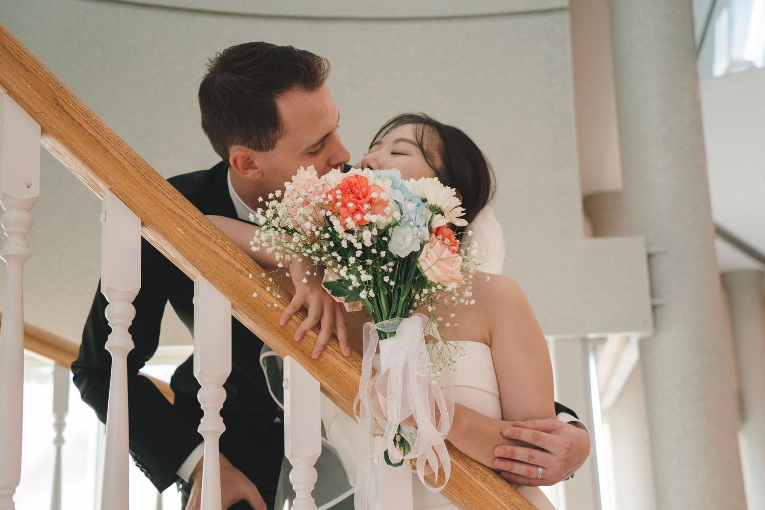 Groom tenderly embraces bride from behind on staircase as she holds a pastel floral bouquet.