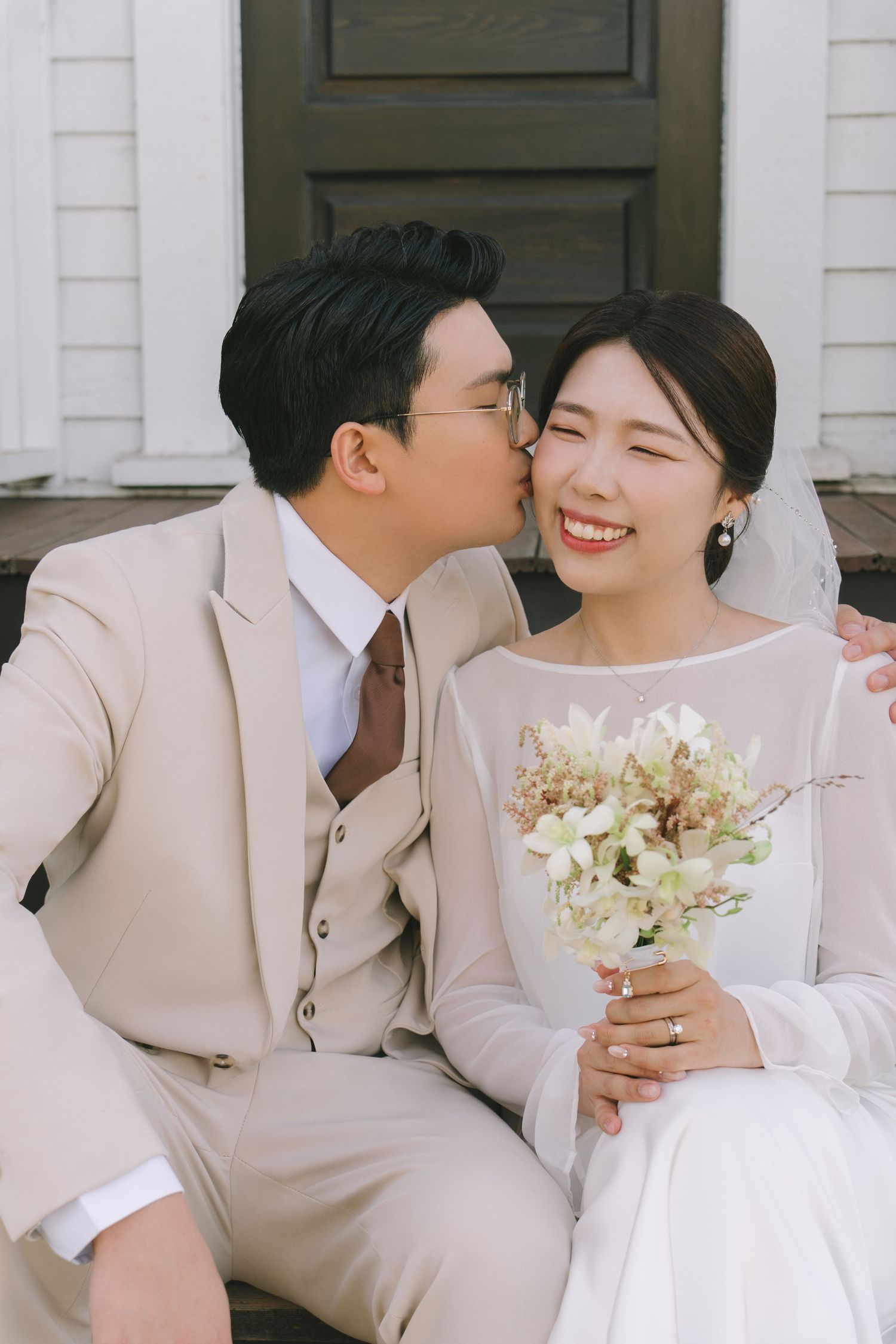 Groom in beige suit kissing bride on cheek as she smiles holding white floral bouquet on church steps.