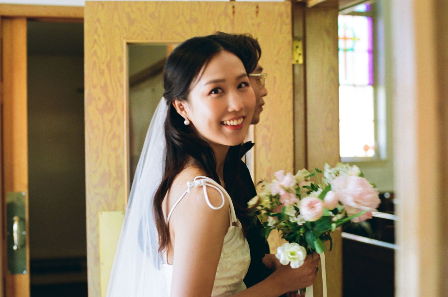 Smiling Asian bride in white dress with veil holds pink bouquet, glancing back over shoulder inside church.