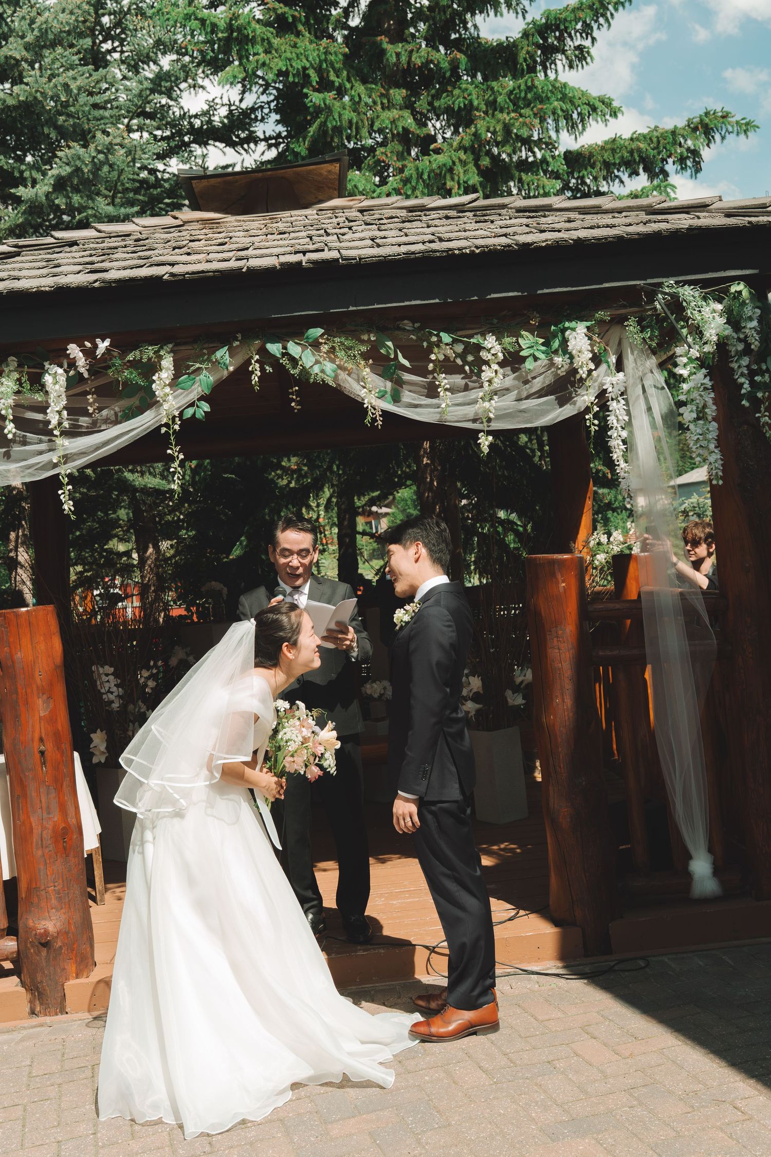 Bride and groom share a joyful moment under a decorated wooden gazebo altar during their outdoor ceremony.