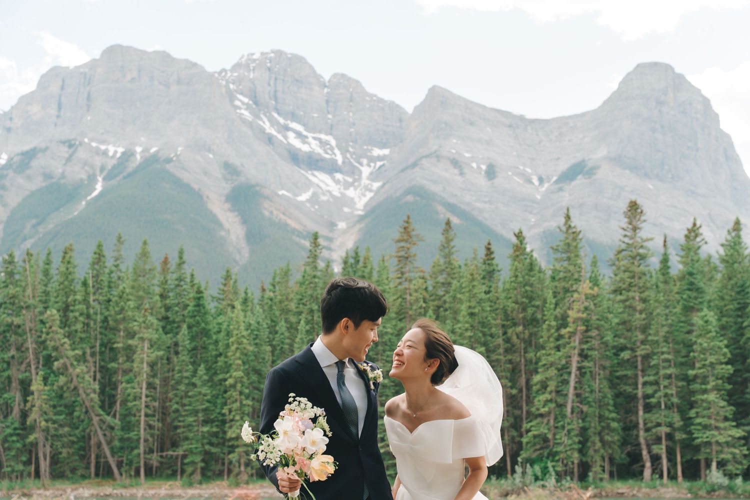 Asian bride and groom gaze at each other lovingly with snow-capped mountains and evergreens behind them.