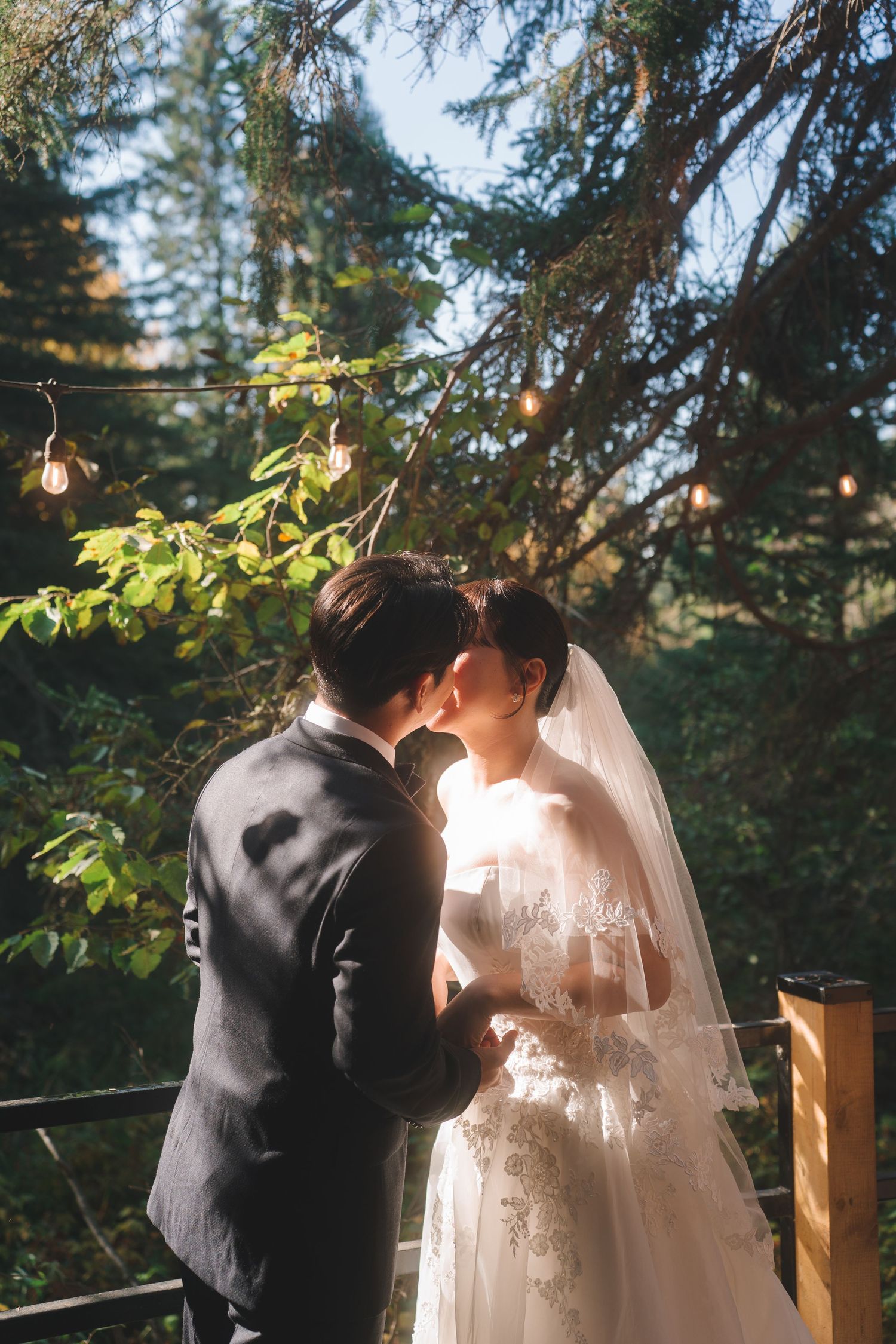 Asian bride and groom share a romantic kiss outdoors surrounded by tall evergreen trees with string lights.