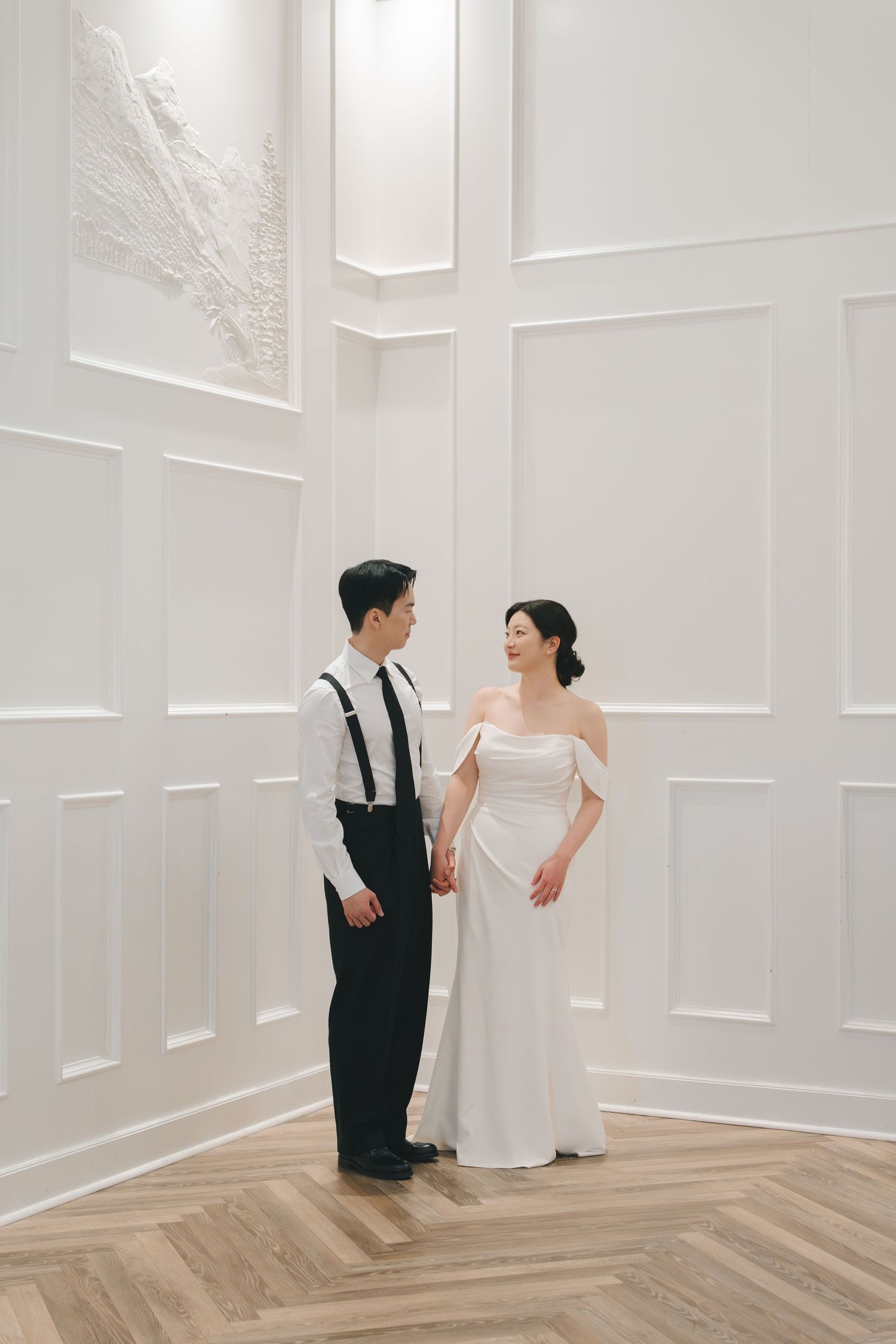 Bride in off-shoulder white gown and groom in suspenders hold hands in bright white paneled room.