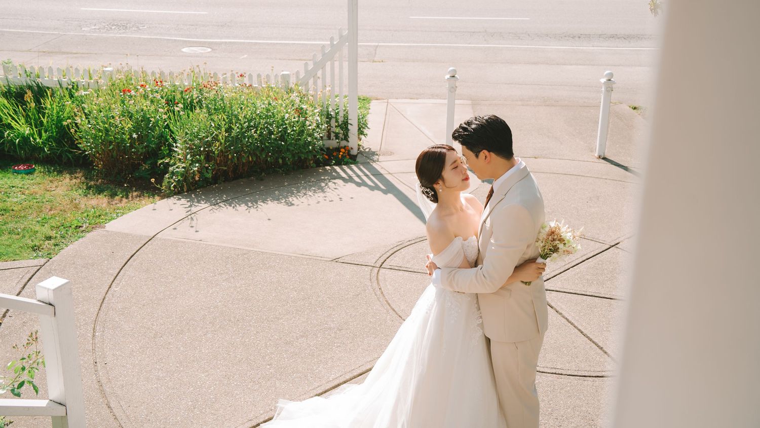 Aerial view of bride and groom embracing near white chapel, bride in flowing white dress holding small bouquet.
