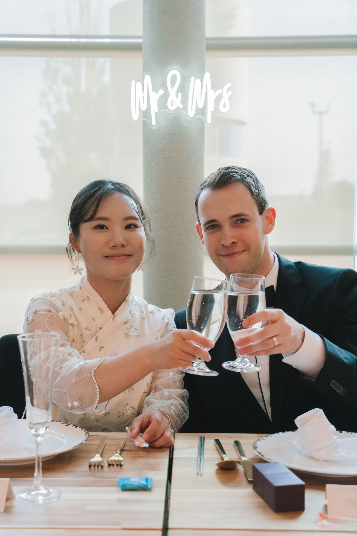 Newlyweds toast with champagne glasses beneath a neon Mr & Mrs sign at their wedding reception dinner.
