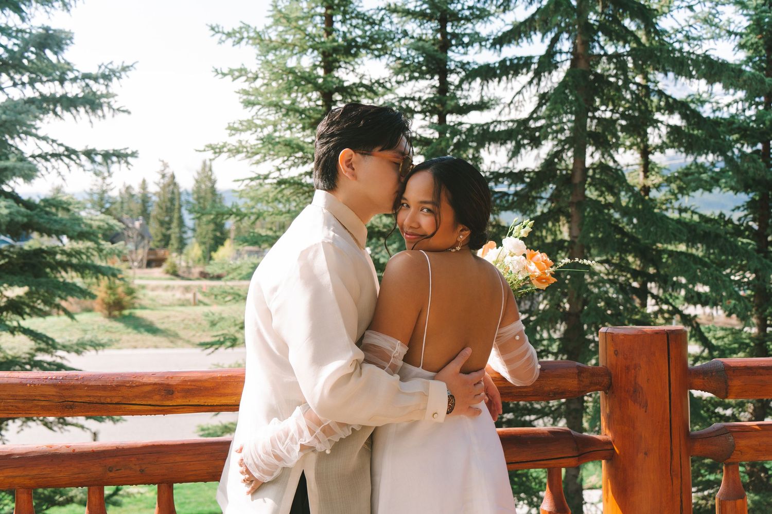 Groom kisses smiling bride on cheek on wooden deck surrounded by lush pine trees outdoors.