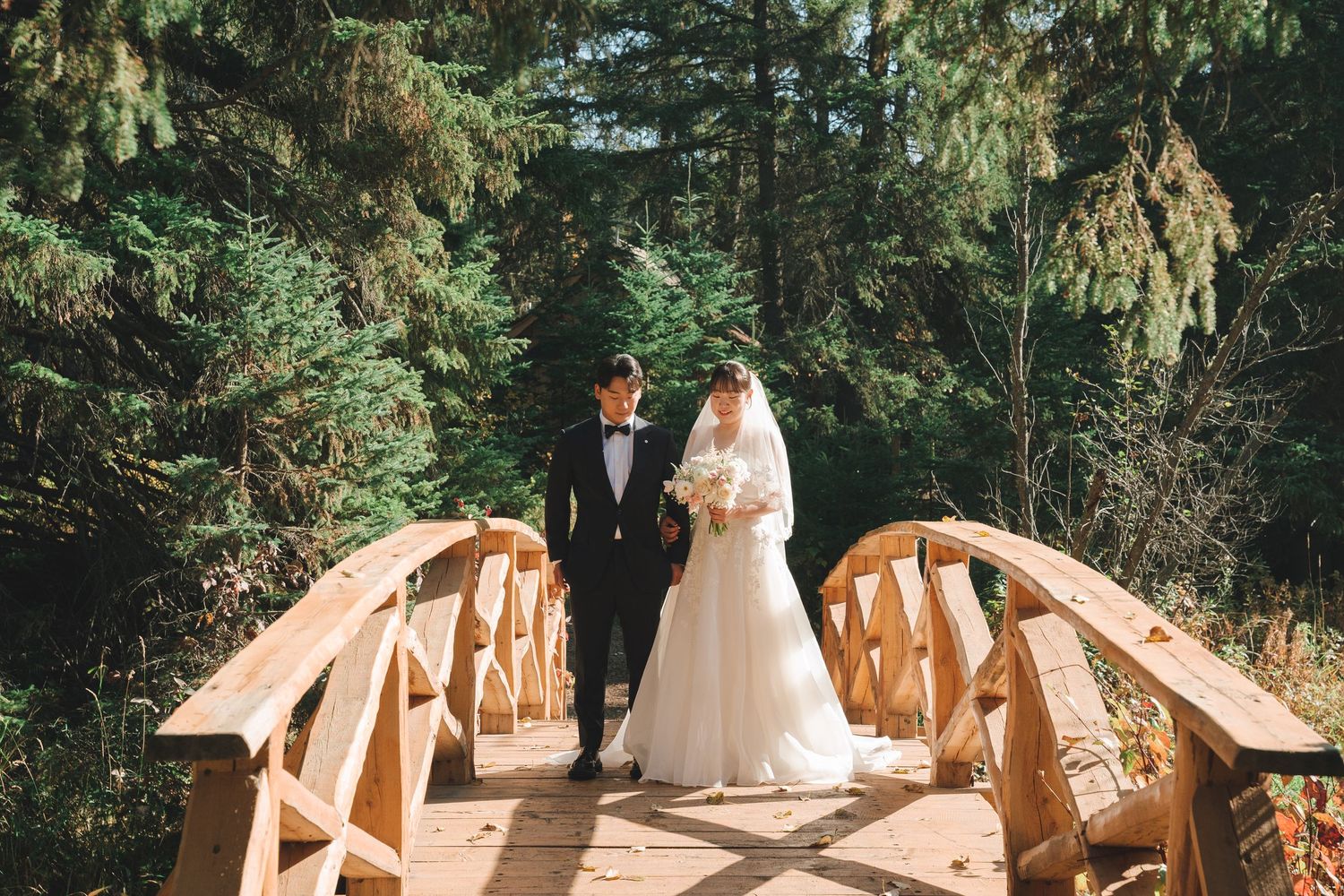Asian bride and groom stand on a wooden arched bridge surrounded by tall evergreen trees.