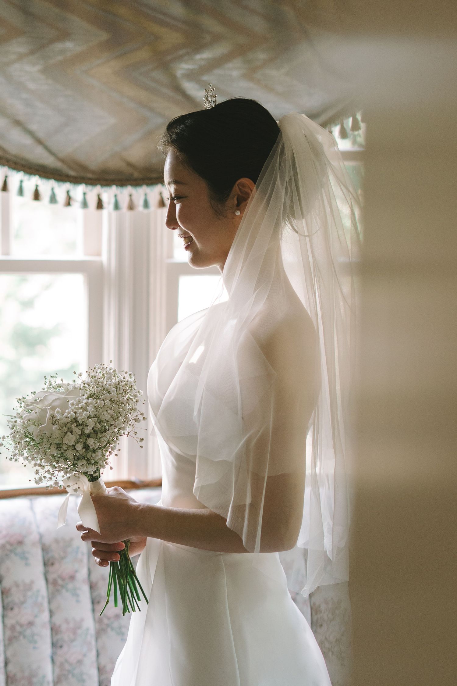 Elegant bride in white gown and veil holds baby's breath bouquet by sunlit window.