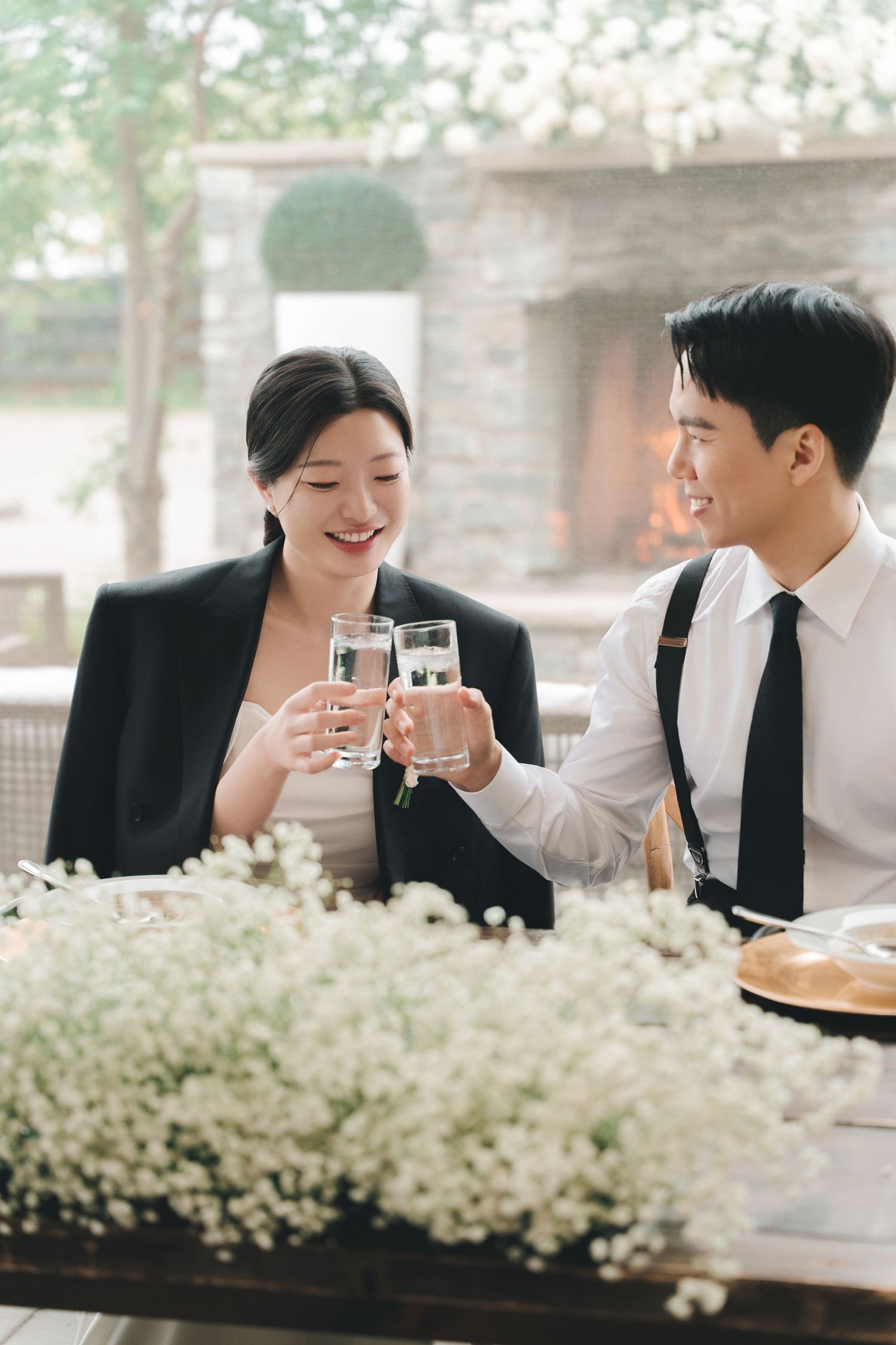Smiling newlyweds in reception attire toast champagne glasses over a table of white baby's breath.