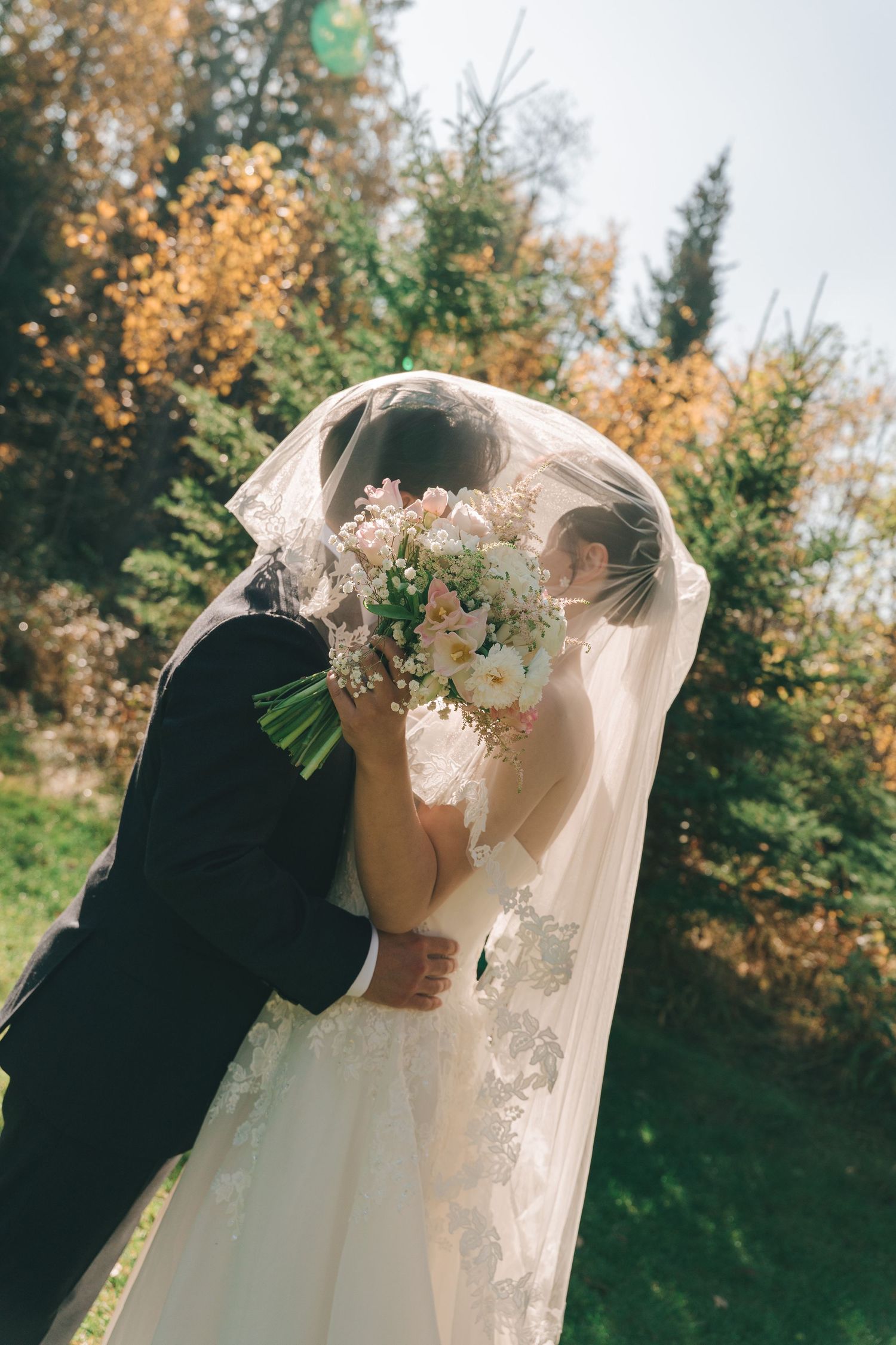 Bride and groom share a kiss under a veil surrounded by autumn foliage outdoors.
