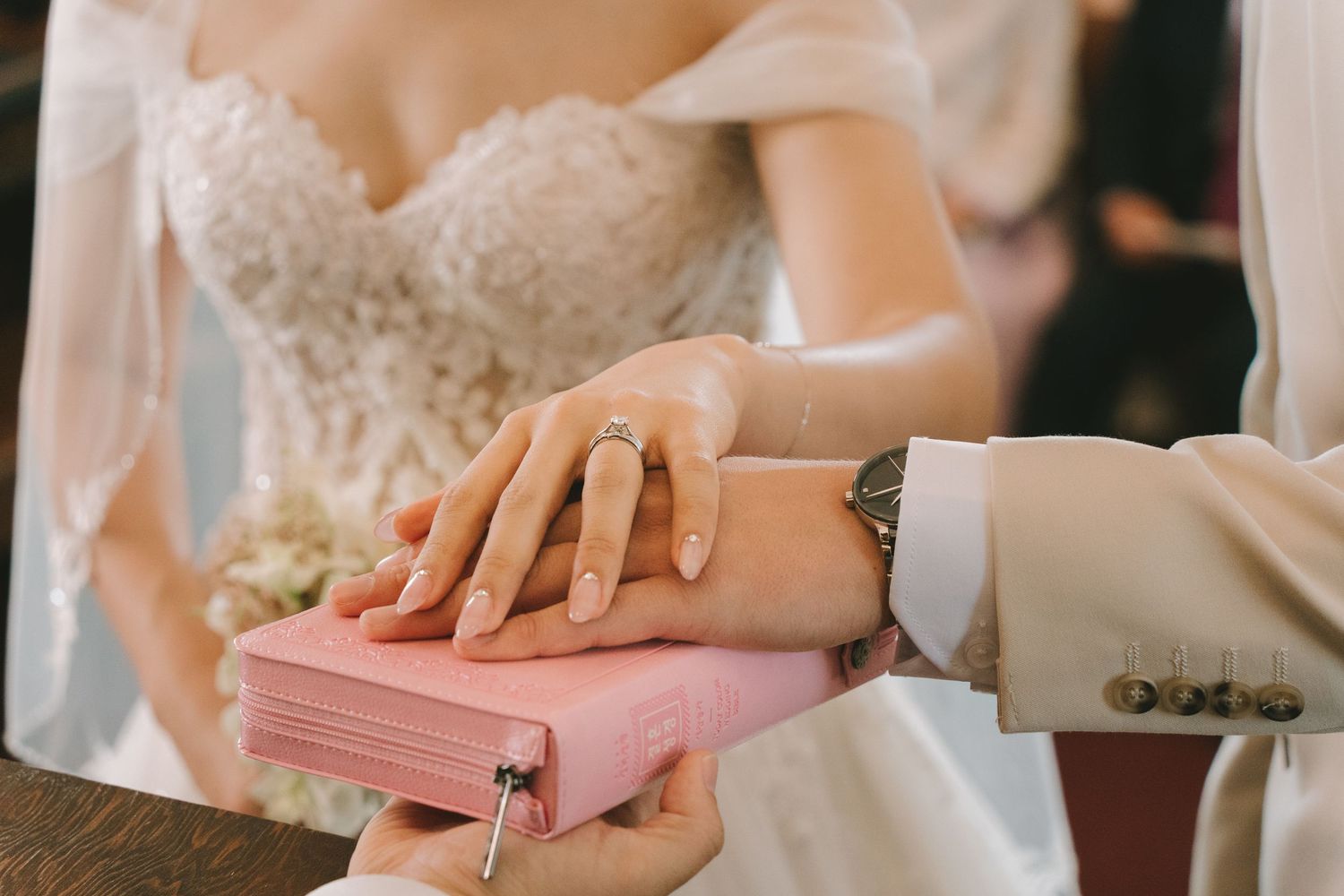 Bride and groom place hands on pink Bible during wedding ceremony, showcasing diamond engagement ring and elegant lace gown.