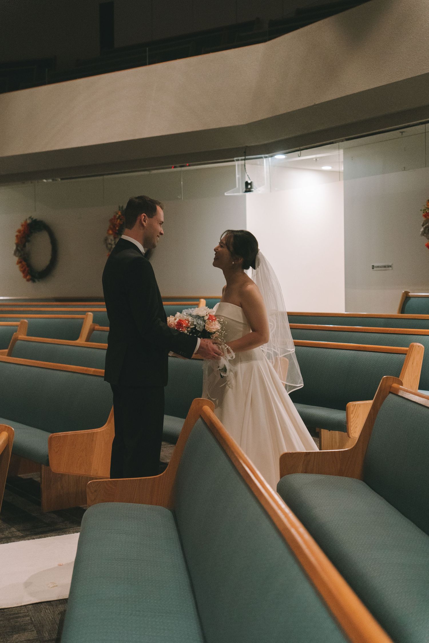 Bride and groom hold hands and face each other between wooden pews inside a church chapel.
