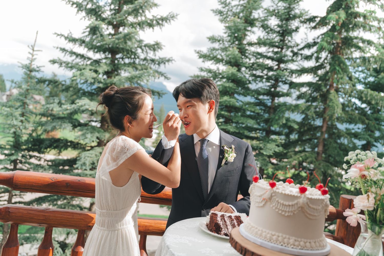 Bride feeding groom cake on outdoor deck surrounded by pine trees.