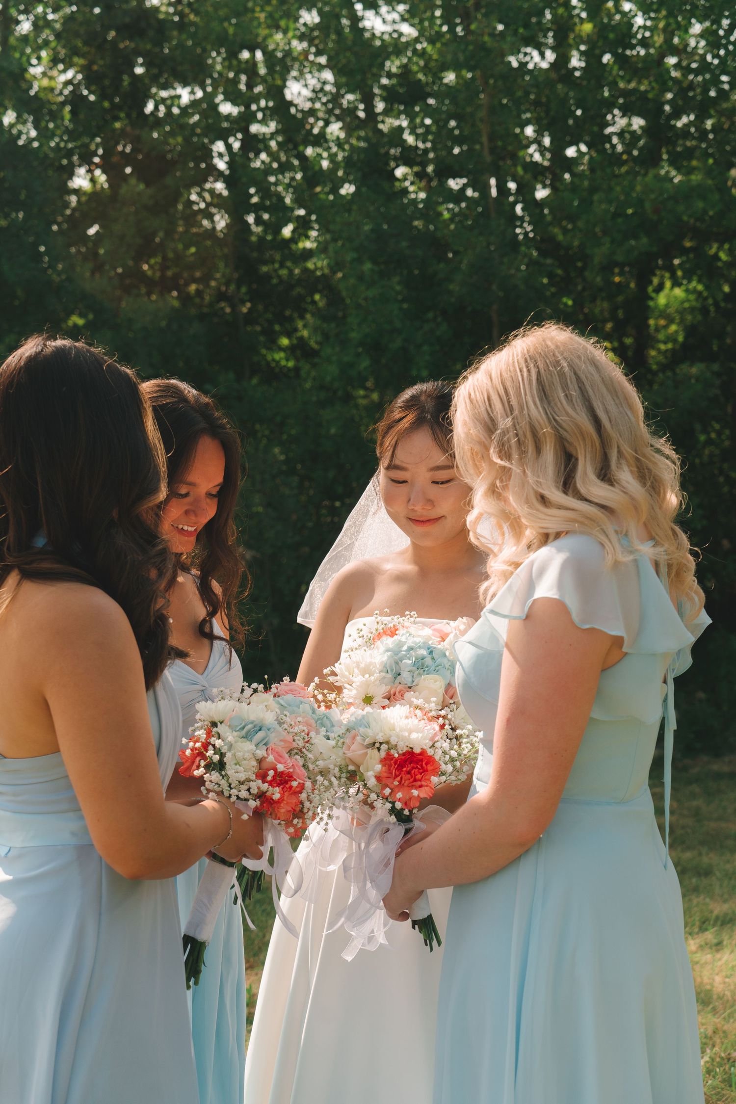 Bride in white gown with veil admires bouquet of pink and white flowers with two bridesmaids in light blue dresses.
