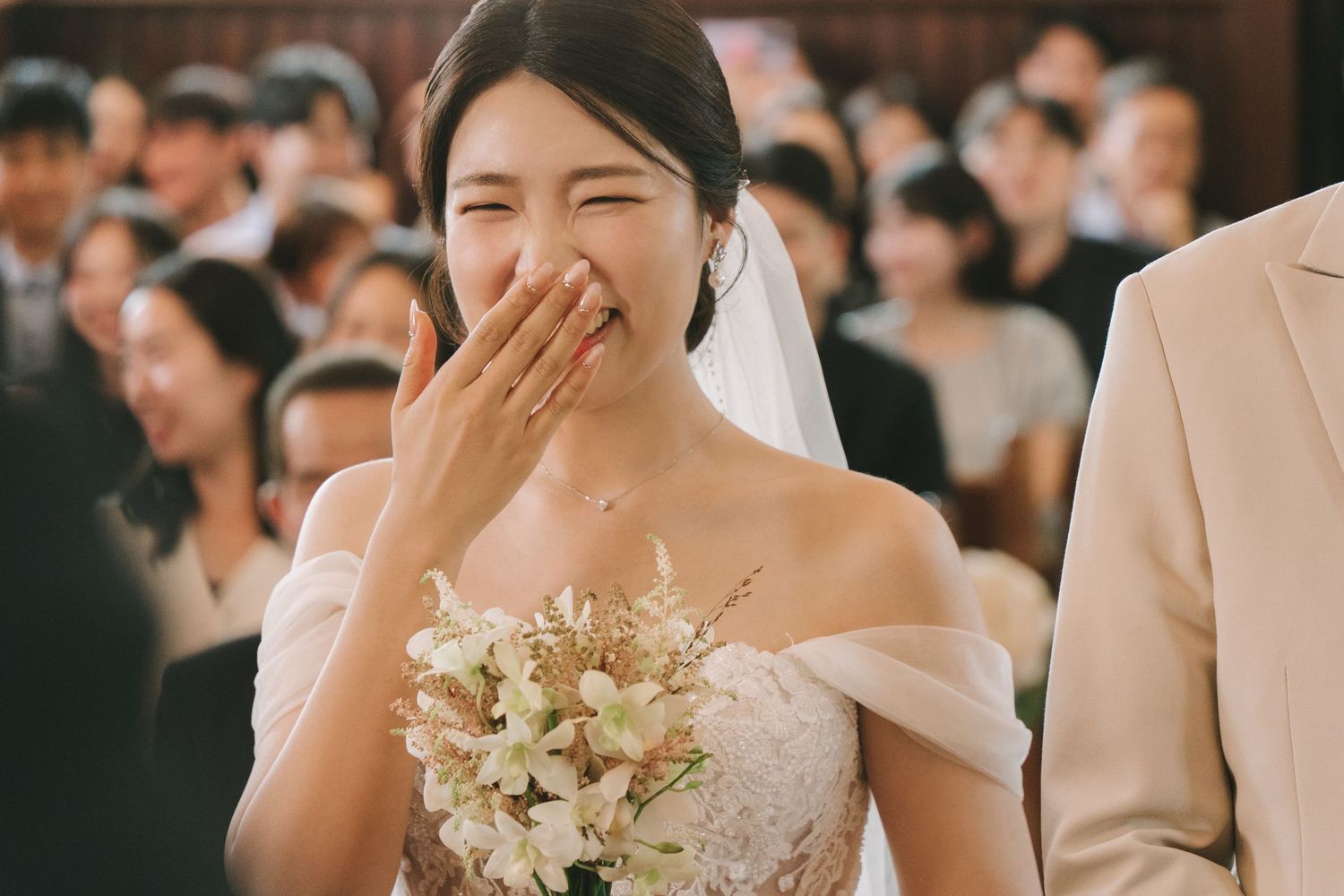 Emotional bride in off-shoulder wedding gown covers mouth while walking down the aisle holding bouquet.
