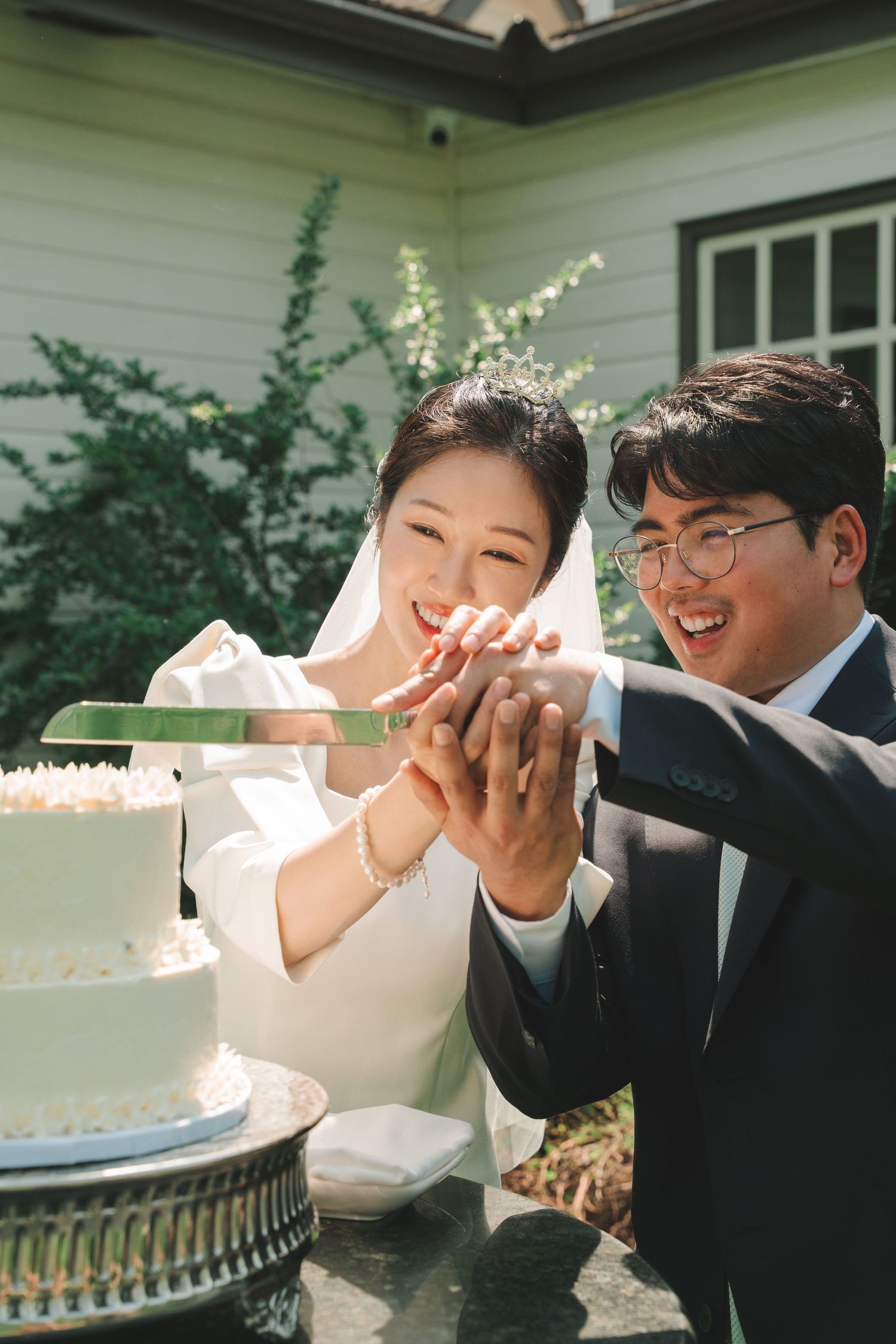 Smiling Asian bride and groom cutting white wedding cake together in sunny outdoor setting.