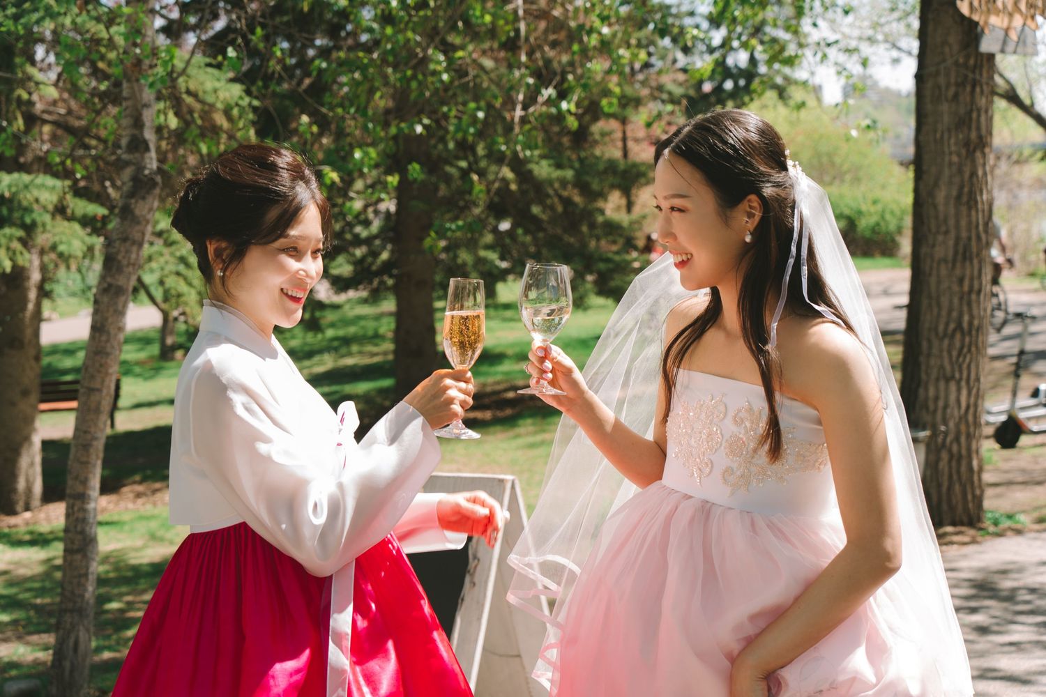 Two women in Korean hanbok and bridal gown toasting with champagne glasses outdoors at a wedding celebration.