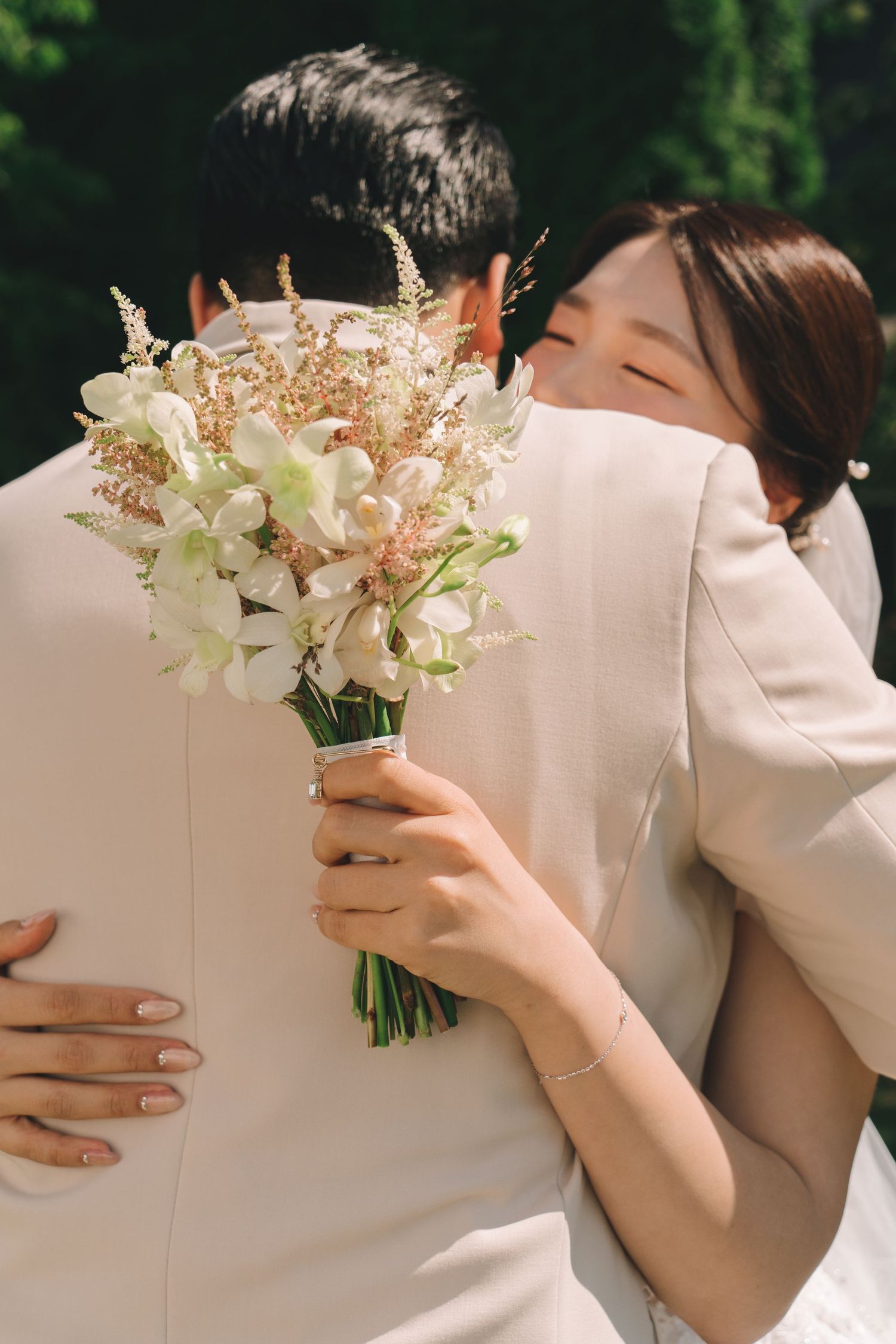 Bride hugging groom from behind holding white floral bouquet outdoors.